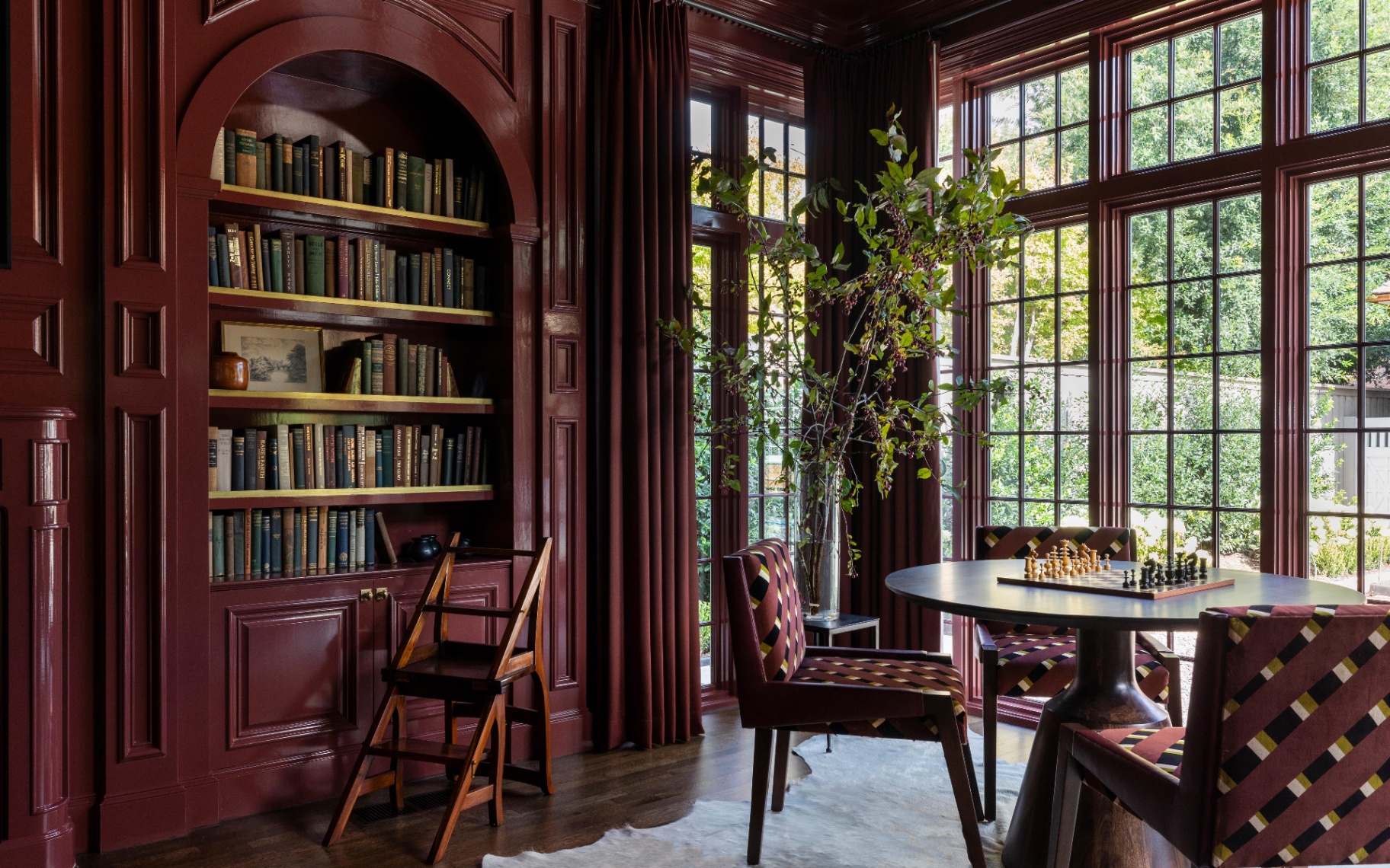 a room with dark red gloss colour drenched walls, shelves, ceiling, window frames and curtains, an arched built in book shelf and a small dining table with a chess board next to a large window