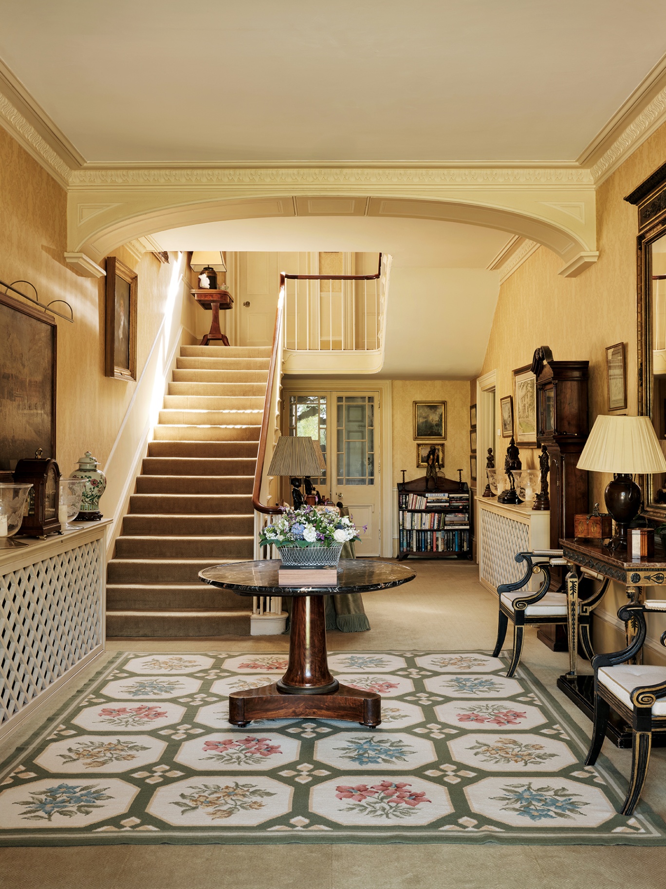 a grand entrance hall in a country house with a large floral rug topped with an antique circular table in the middle and a large staircase on the left. To the right are a pair of antique wooden chairs and sideboard.