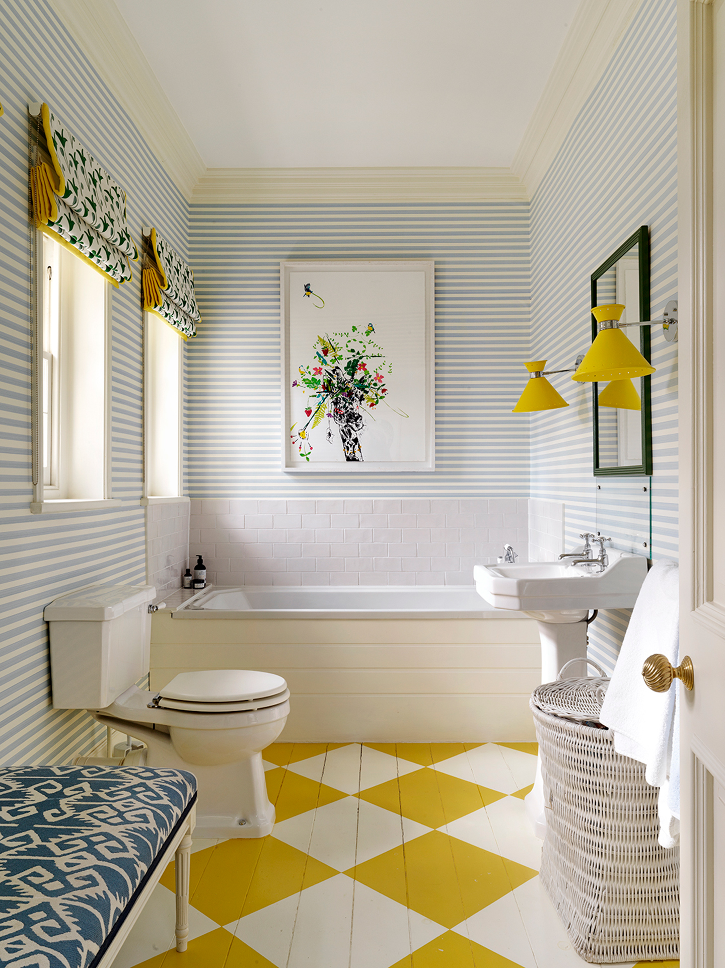 A colourful bathroom with blue and white striped wallpaper and yellow and white painted checkerboard flooring. A bath is built in at the far end, with a toilet and sink in front and a bench and laundry basket in the foreground