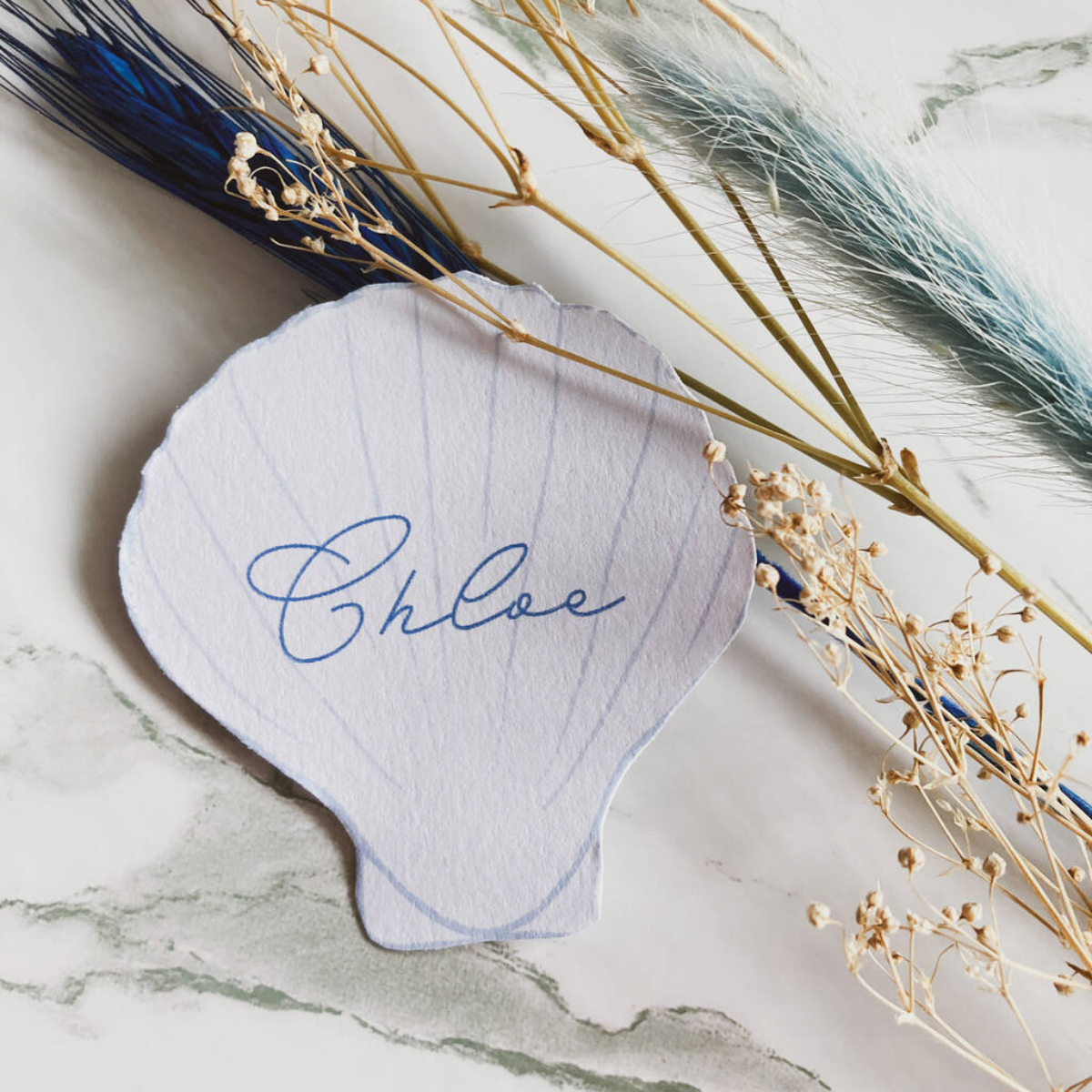 a white and blue seashell shaped place card with the name chloe in cursive writing with sprigs of neutral and blue dried pampas