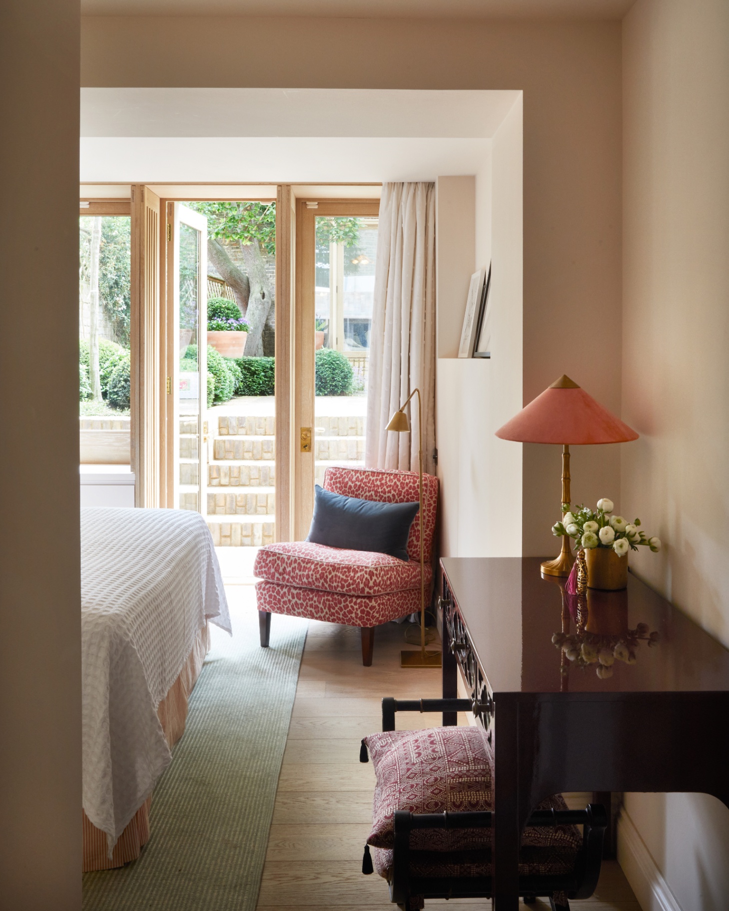a bedroom with cream walls, a pink patterned accent chair with a gold floor lamp and an antique dressing table