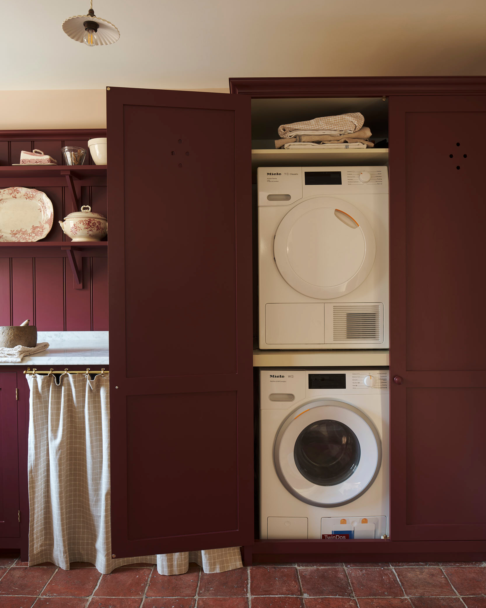 a utility room with orange stone floors, dark red cabinetry and a washing machine and a tumble dryer stacked on top of eachother
