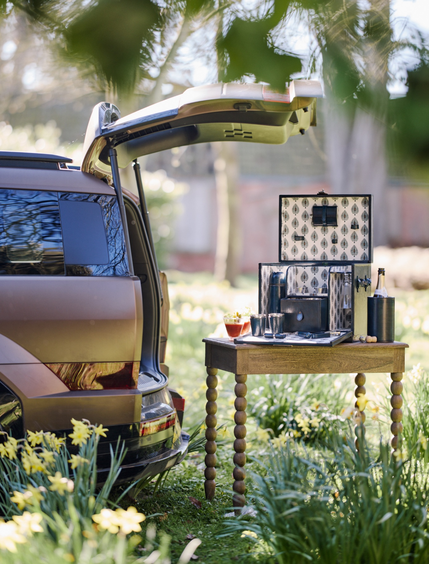 A CAR WITH AN OPEN BOOT AND A BOBBIN WOOEDN OUTDOOR TABLE WITH A NAVY LEATHER PORTABLE DRINKS BAR