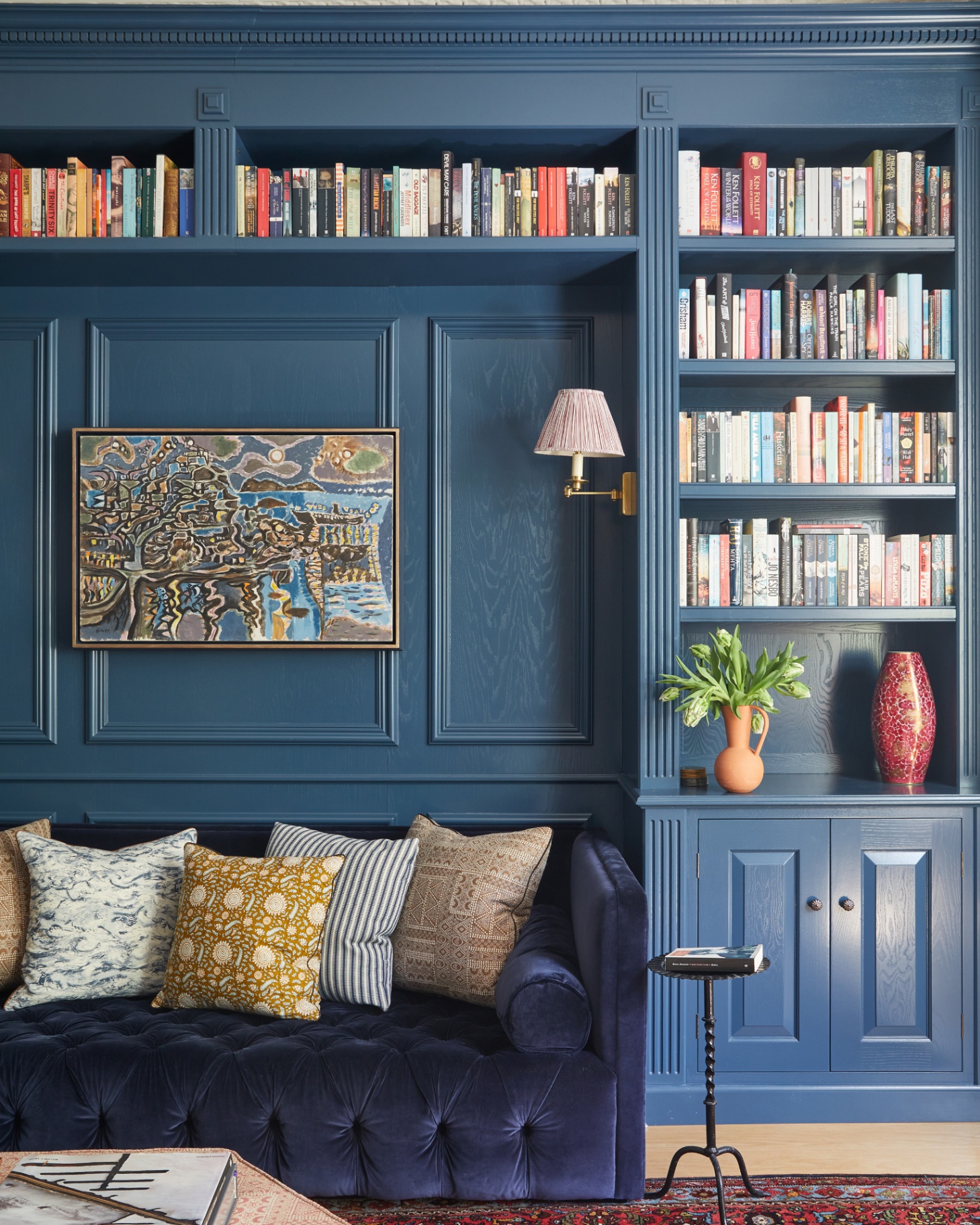 a built in bookcase painted in dark blue and filled with colourful books and a dark navy velvet sofa with blue adn yellow cushions