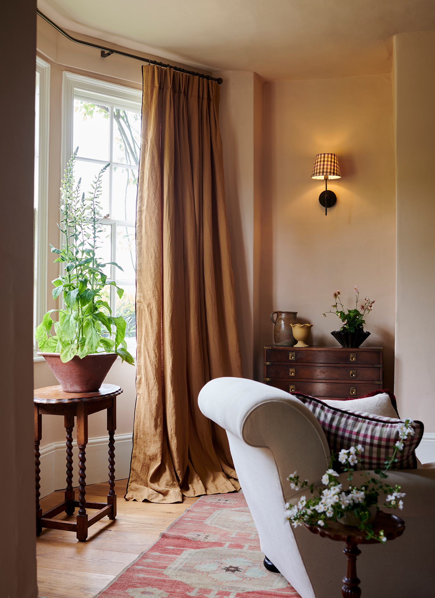 a living room with long ochre curtains, a dark wooden vintage chest of drawers with vases on top and a small wooden table in front of the window with a plant on top