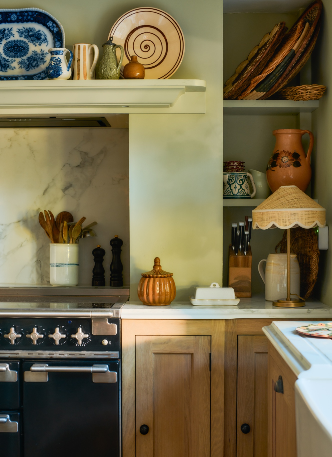 A closeup of a kitchen corner with sage green walls, a shelf above the range alcove with plates and built in alcove shelves next to it