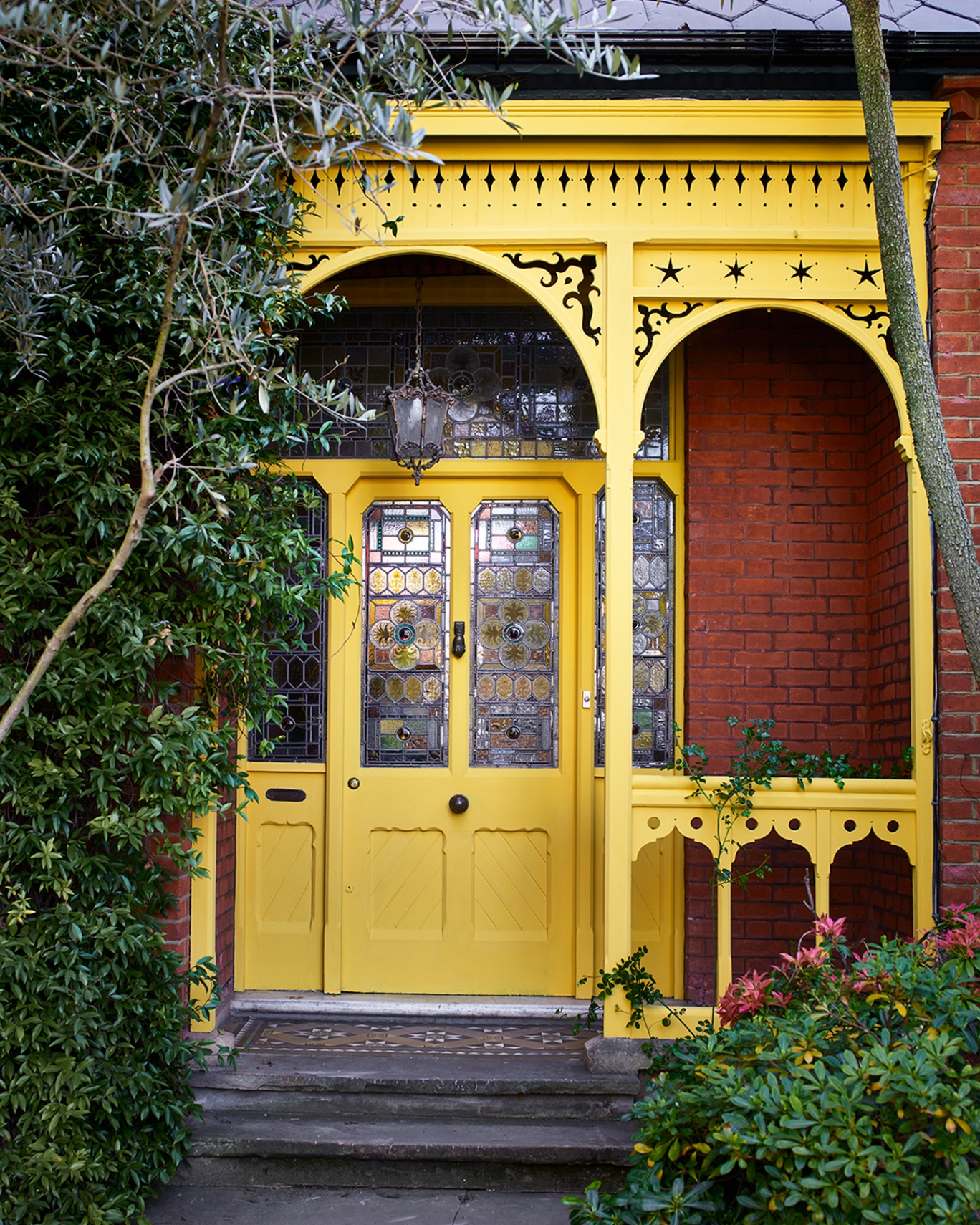 a bright yellow front door, a yellow porch with a lantern pendant, stained glass panels, steps and shurbs either side of the path