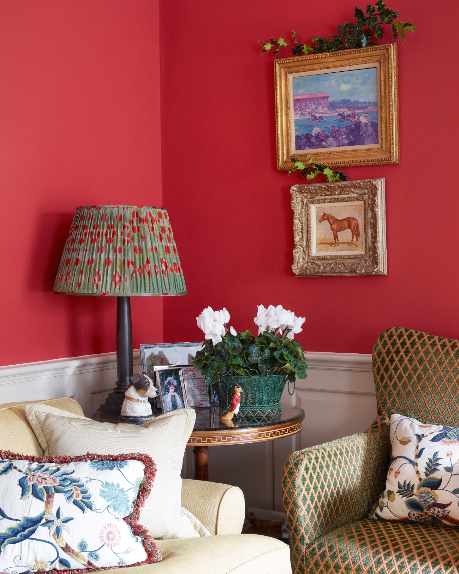 a living room corner with bright red walls, two gold framed prints hanging above a green and beige patterned armchair and a side table with a green and red pleated lamp