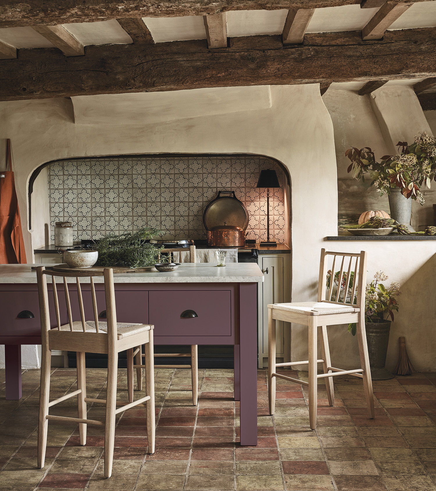a country kitchen with wooden beams, a range cooker in a nook, flagstone flooring, a purple kitchen island and light wood chairs