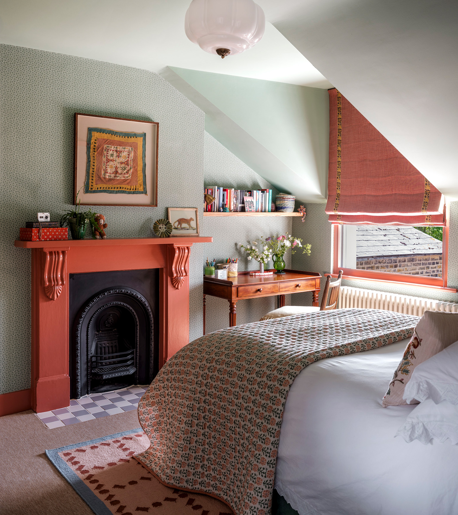 a bedroom with green sloping ceilings, coral pink blinds on the window, a coral pink painted fireplace with art hanging above it and a bed with a knit throw on the end