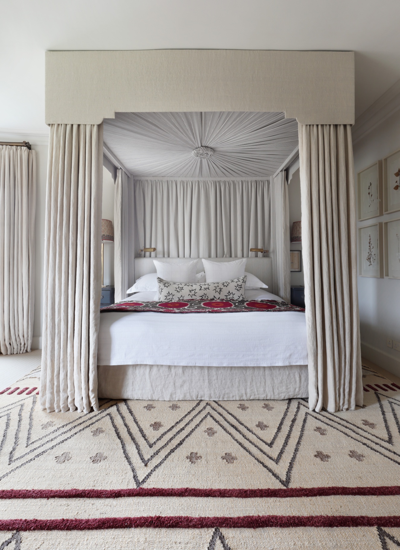 An luxurious, serene bedroom featuring an upholstered four-poster canopy bed draped in tailored neutral linen fabric, set over a patterned cream and maroon geometric rug.