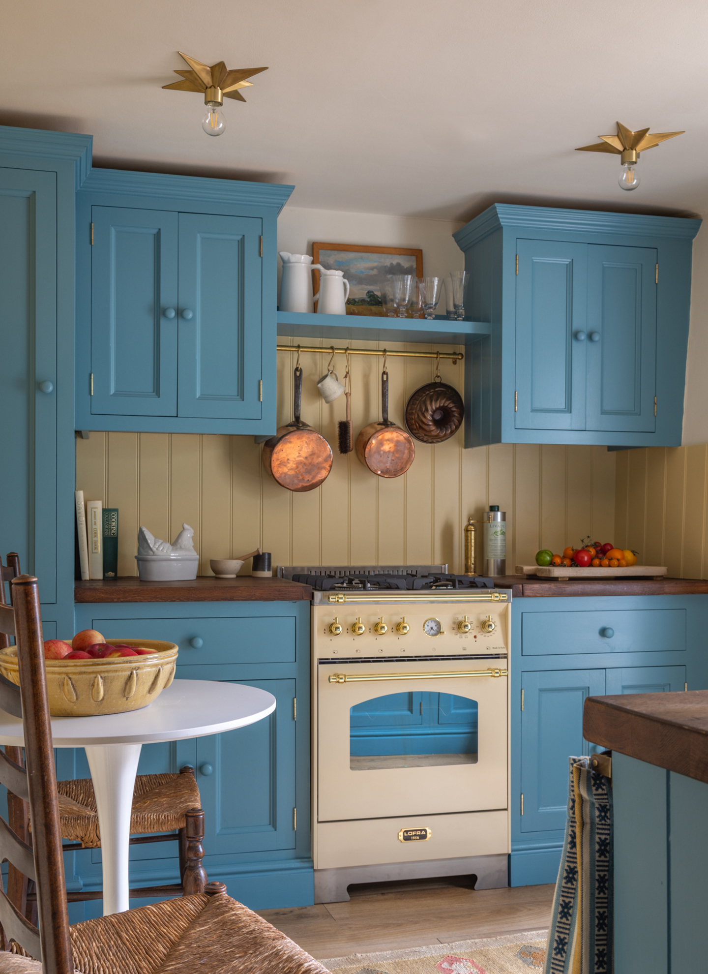 a small kitchen with bright blue cabinetry, a cream range cooker with copper pans hanging above it and two star shaped lights on the ceiling