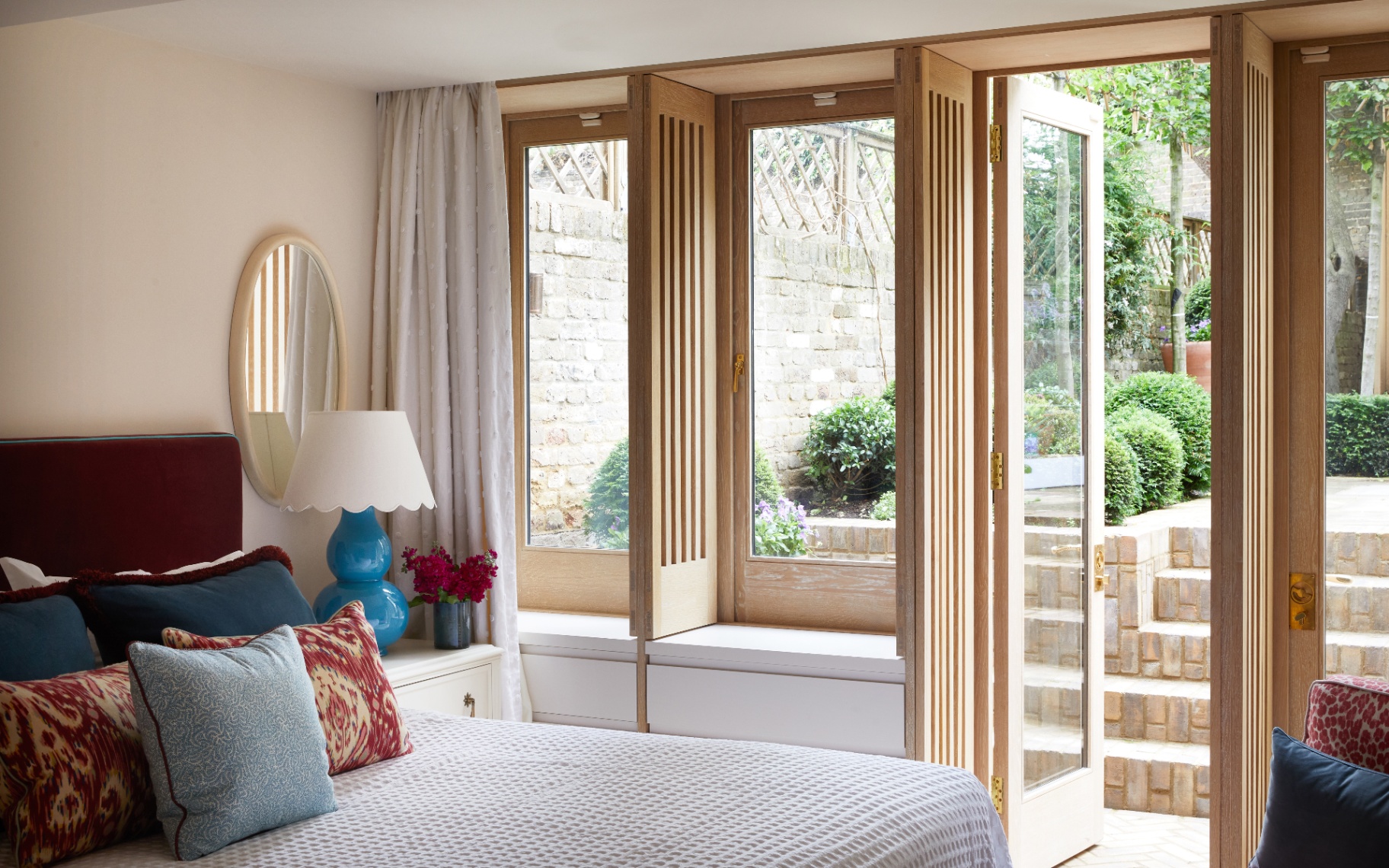 a bedroom with large wooden windows open onto the garden and a bed with white bedding and colourful cushions