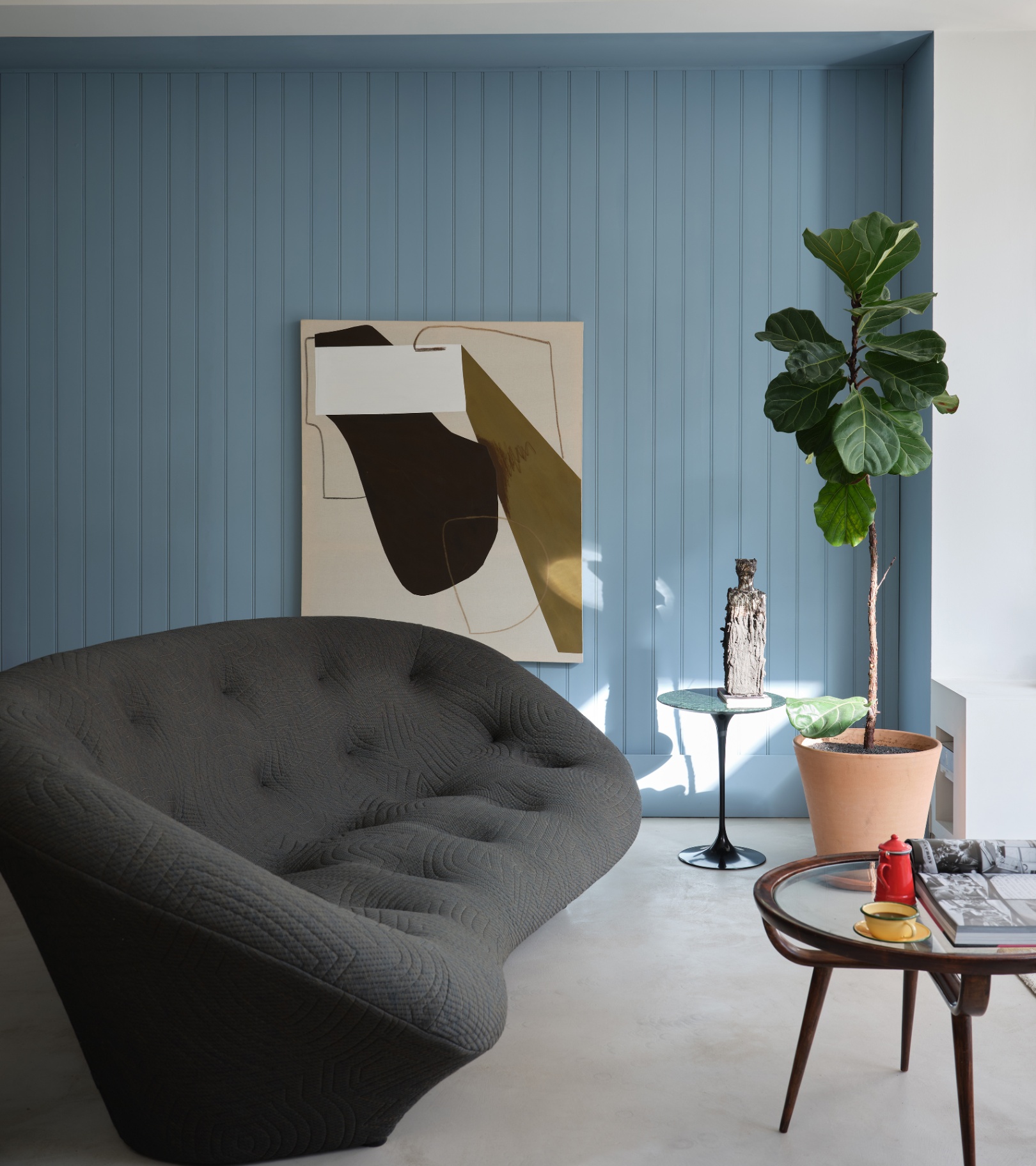a living room with blue painted paneled alcove and an abstract painting hung on it, a large modern style charcoal sofa and a large plant in the corner
