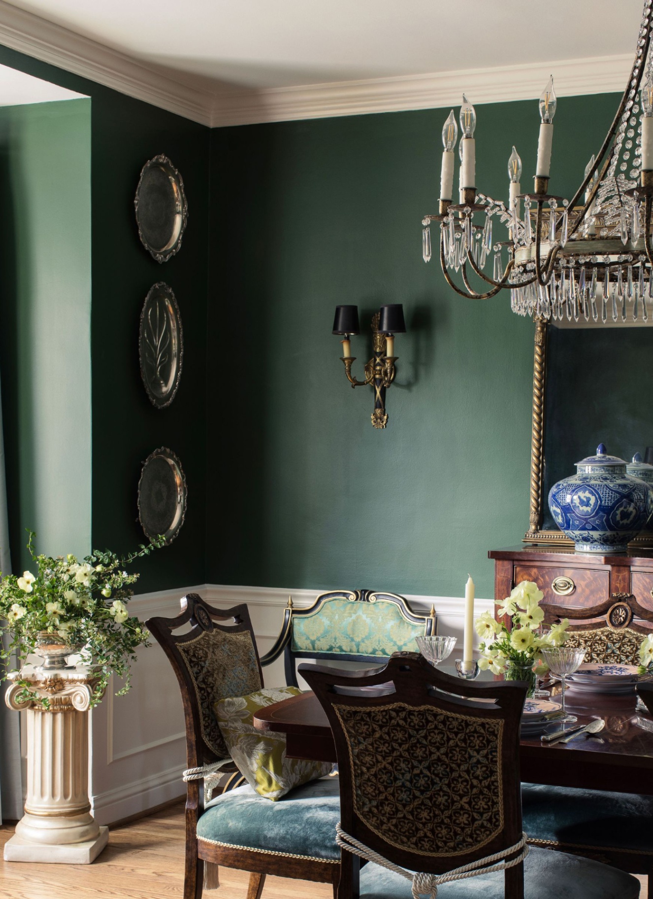 Elegant emerald green dining room featuring white wainscoting, a crystal chandelier, dark wooden dining table set with silver and blue ceramics, and silver platters mounted on the wall.
