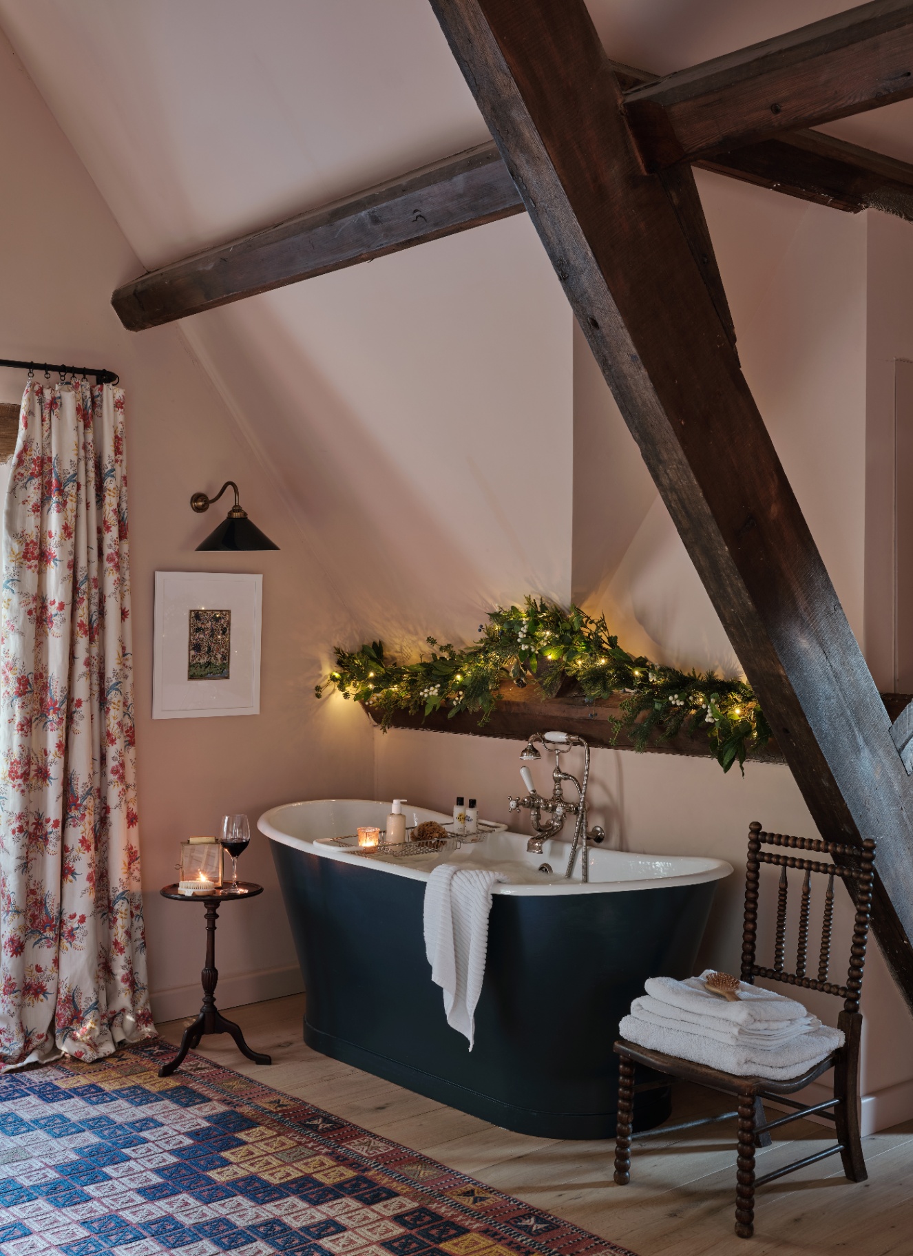 A freestanding roll-top bath beneath a dark wooden beam decorated with a fairy-lit green garland in a cozy bathroom