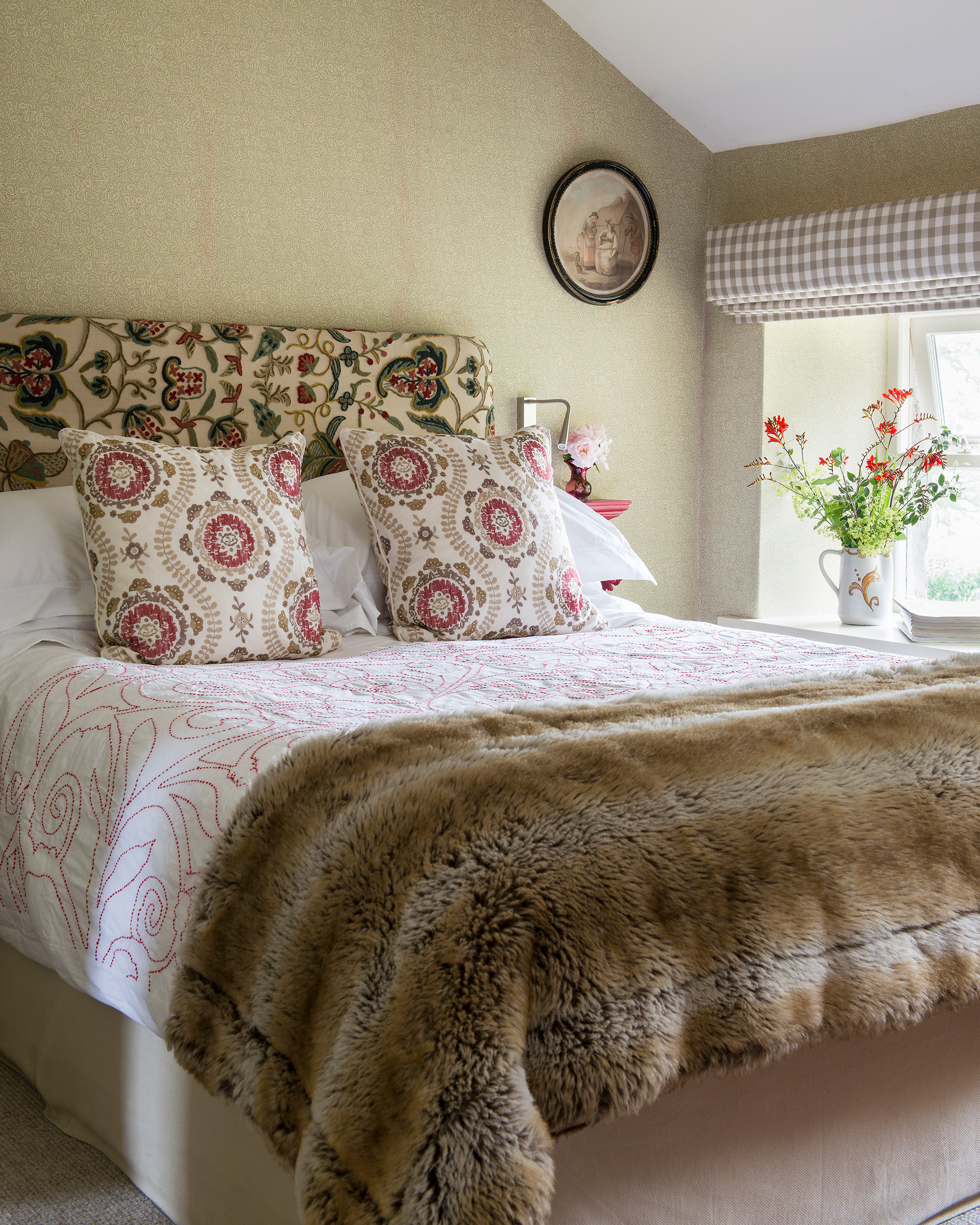 a bedroom with yellow wallpaper, a bed with a cream and red floral headboard, white and red floral cushions and a faux fur throw