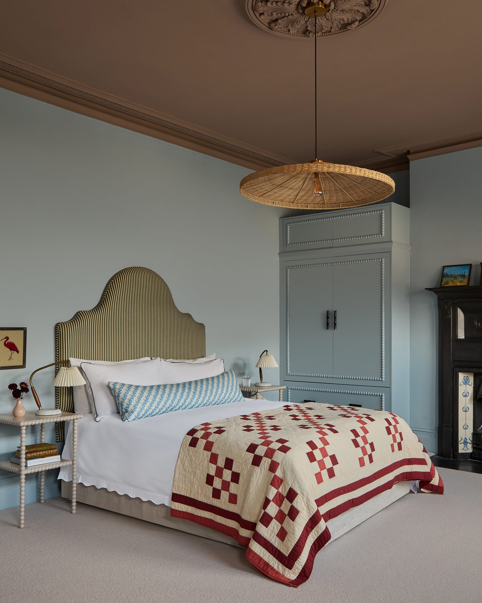 a bedroom with pale blue walls, a brown ceiling, a rattan pendant light, a double bed with a green and white stripe headboard and a red patterned bedspread and a white bobbin bedside table