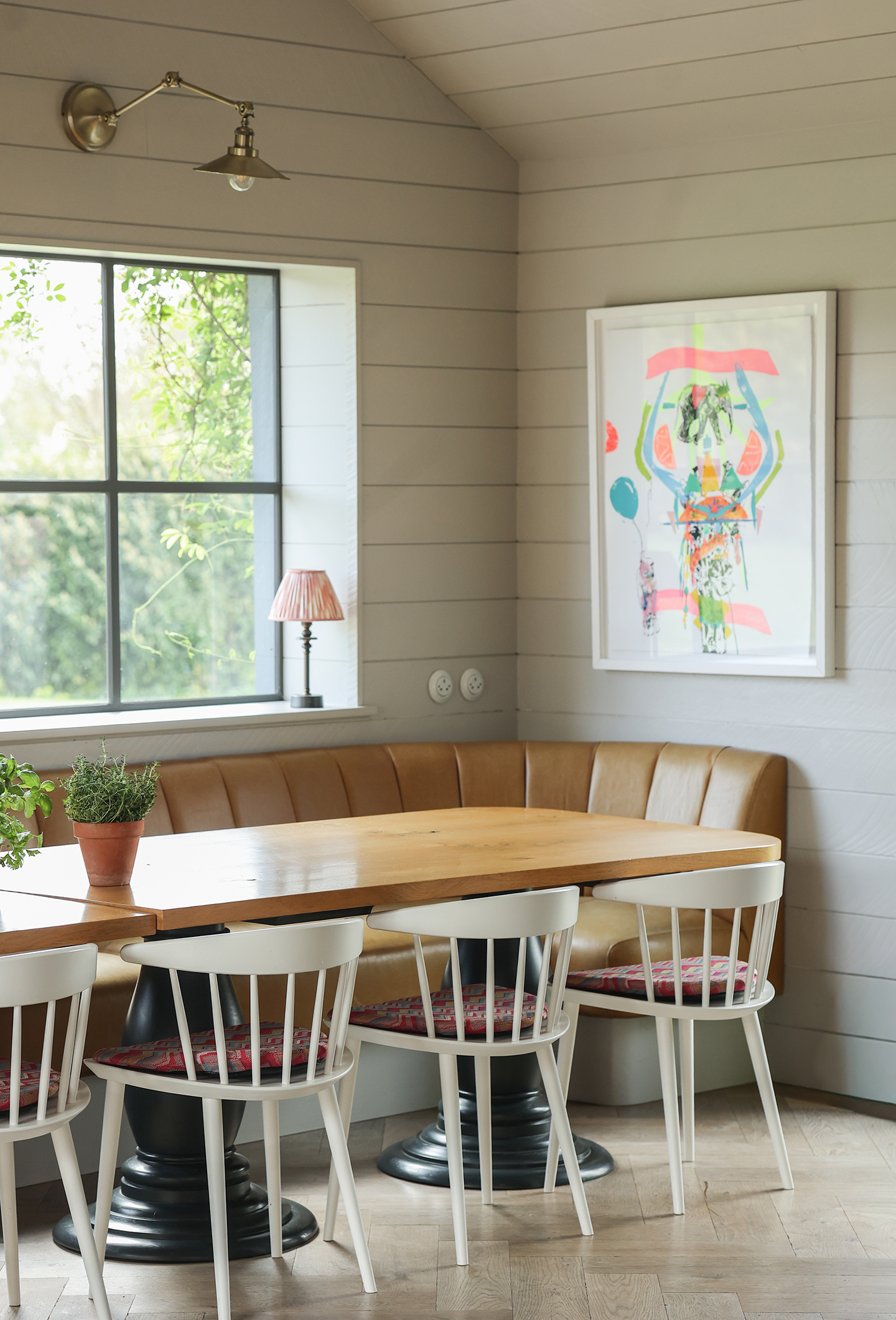 A close up of a dining area in a kitchen with tongue and groove walls painted in a neutral shade. A wooden dining table is paired with white chairs with red seat cushions and leather banquette seating. A large abstract art hangs on the wall above