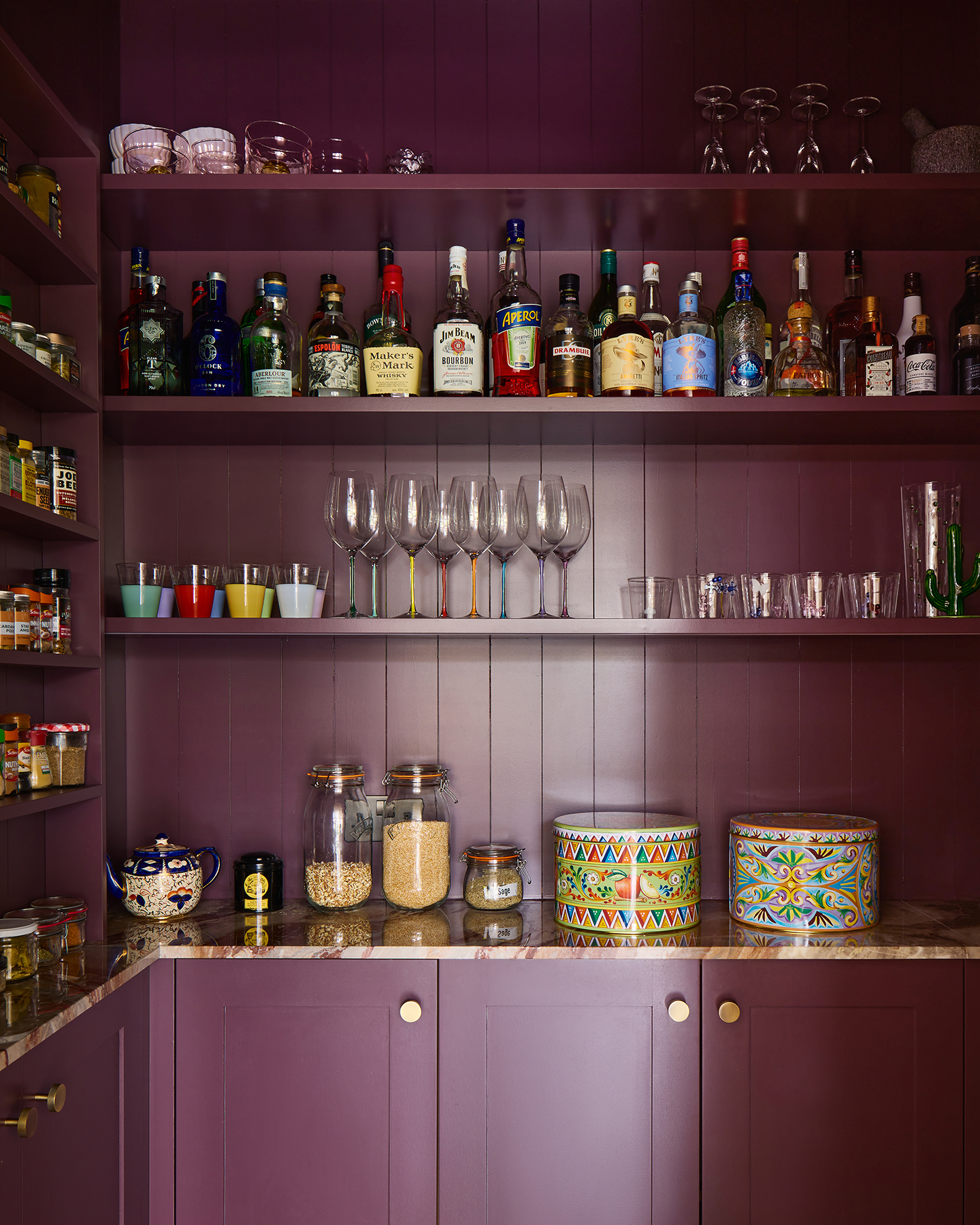 A dark purple pantry with tongue and groove walls and marble worktops on the lower cabinets. Open shelving holds bottles, glasses, tins and jars 