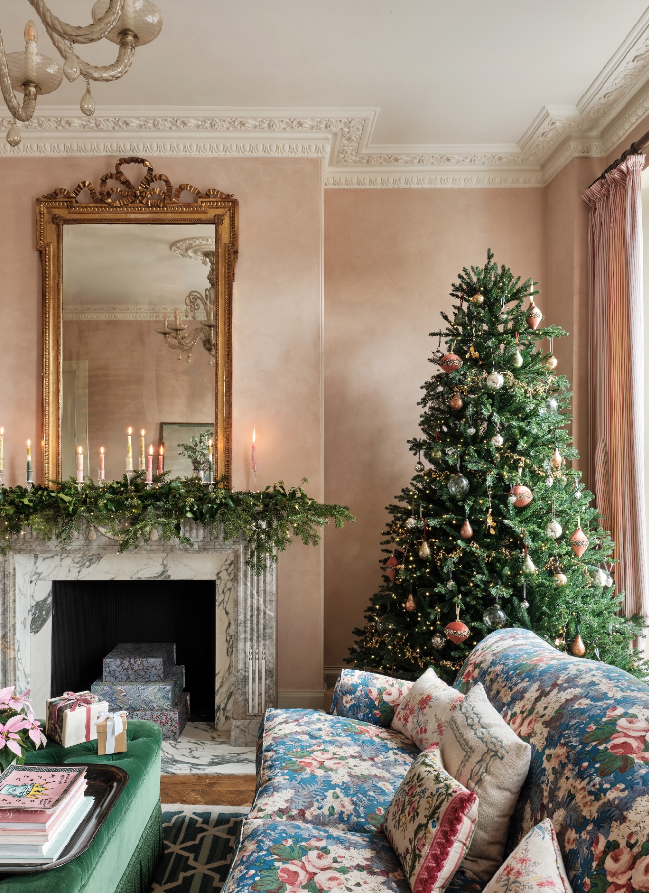 A decorated Christmas tree with pink and glass baubles next to a marble fireplace adorned with a green garland and pastel candles in a blush pink living room