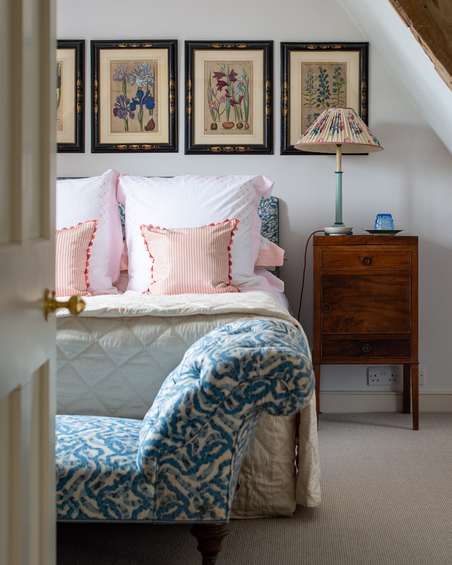 a bedroom with sloped ceiling and wooden beams, a double bed with a set of 4 vintage botanical art prints above it and a blue and white patterned bench at the foot of the bed