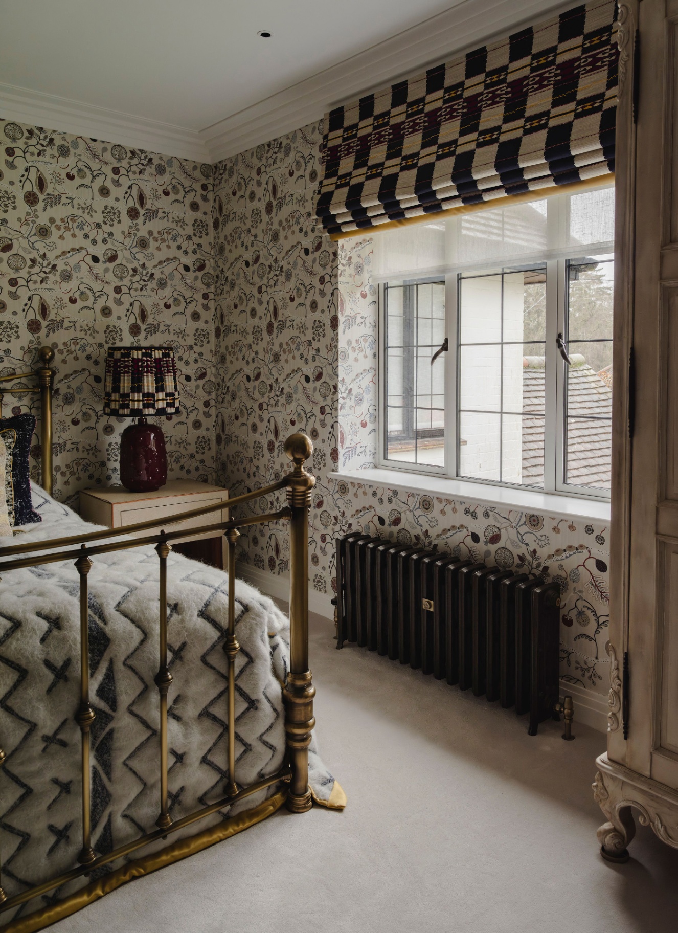 a bedroom corner with white and floral patterned wallpaper, a window with a low black radiator underneath, a gold iron framed bed and a white bedside table with a dark red lamp with a white and black shade