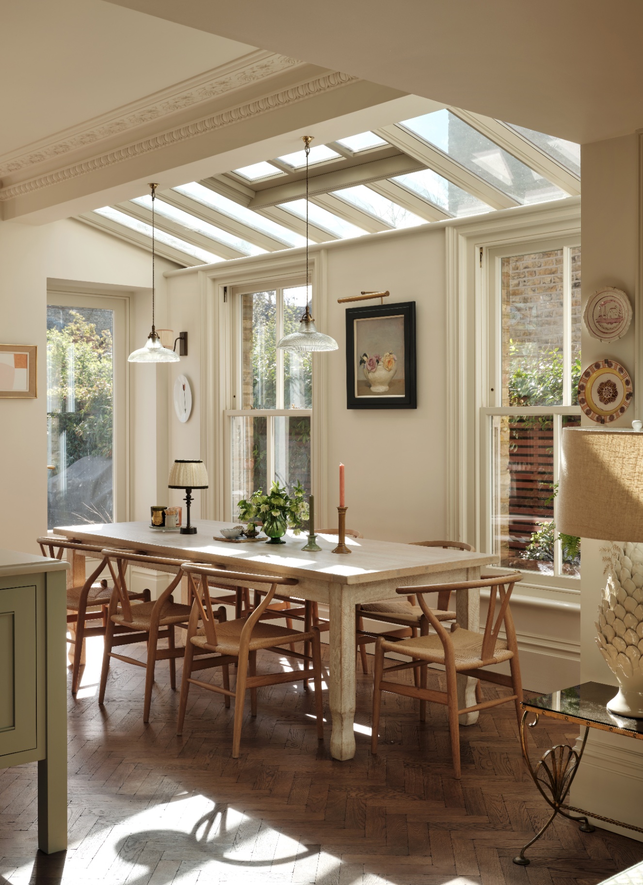 Sunlit dining space under a glass roof lantern featuring a long wooden table and twin fluted glass pendant lights.