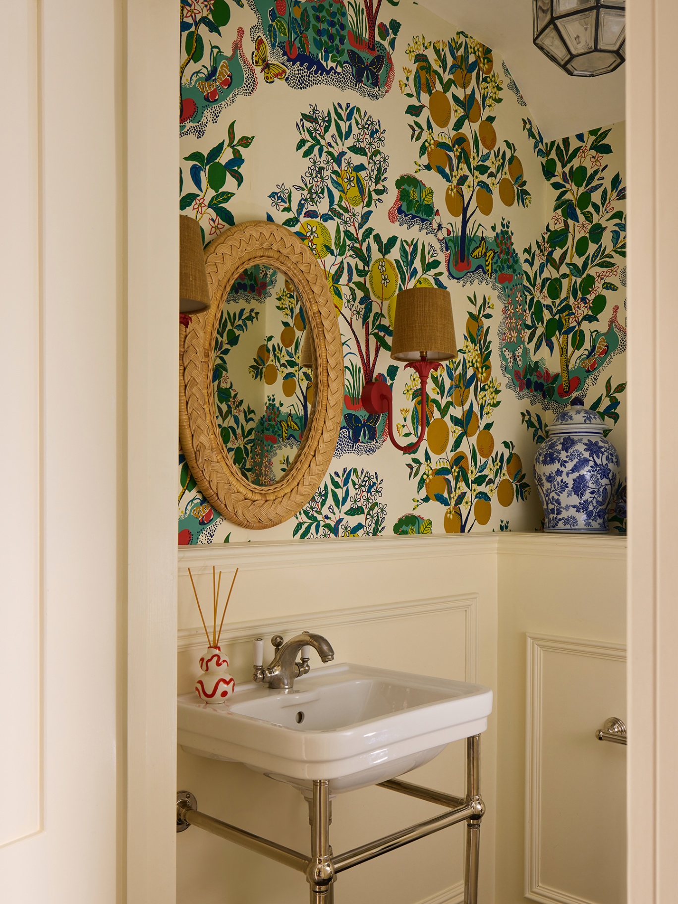 A close up shot looking through a doorway into a cloakroom with panelled cream lower walls and a botanical wallpaper on the upper half of the walls. There is a classic basin and wall lights and a rattan mirror hang on the wall