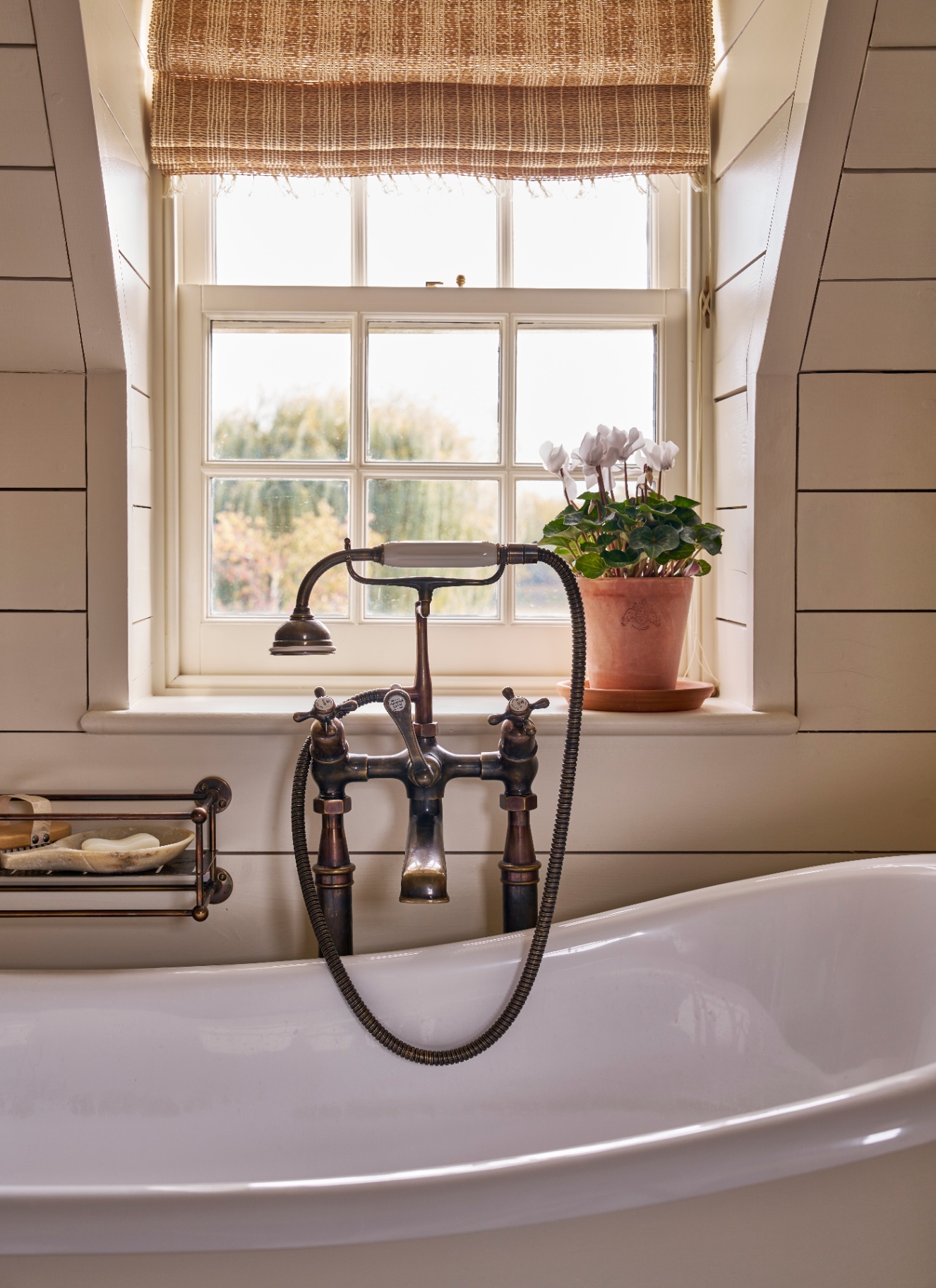 a close up of a freestanding bath with a vintage tap and shower head in front of a window with a blind and a terracotta flower pot