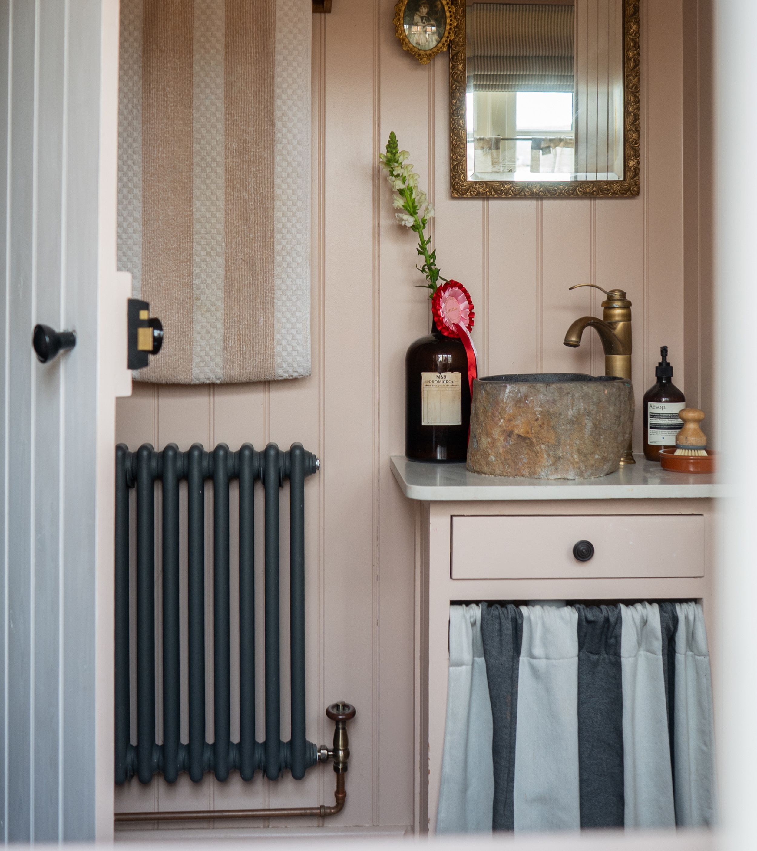 tori murphy small bathroom with plaster pink panelled walls, a marble sink with a blue and navy striped skirt undernearth, a gold tap and a gold mirror and a navy radiator