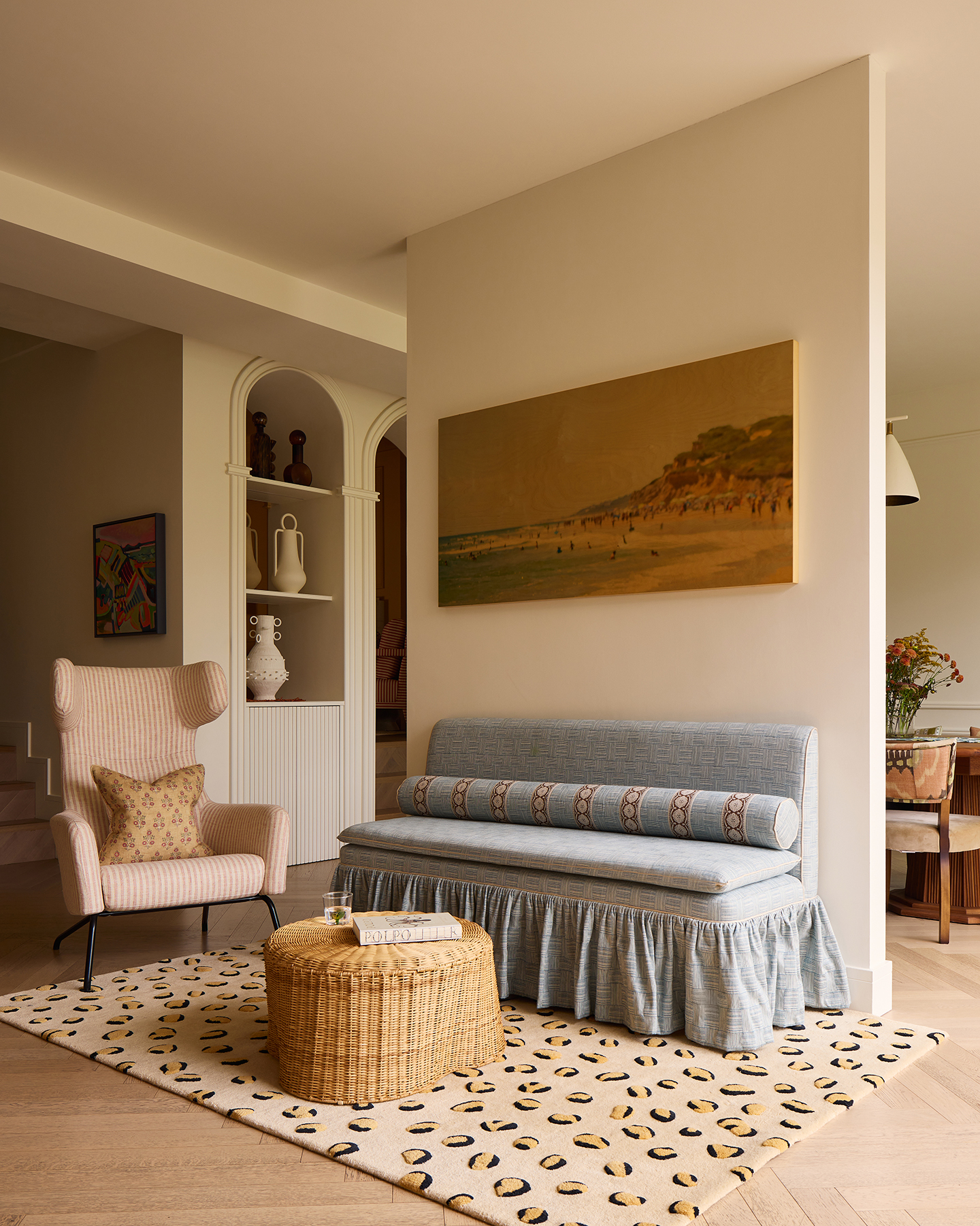 An upholstered skirted bench with a long bolster cushion sits against a partition wall, with a leopard print rug below and a striped armchair to the side. A small rattan table sits in the centre. Arched built-in alcoves can be seen in the background