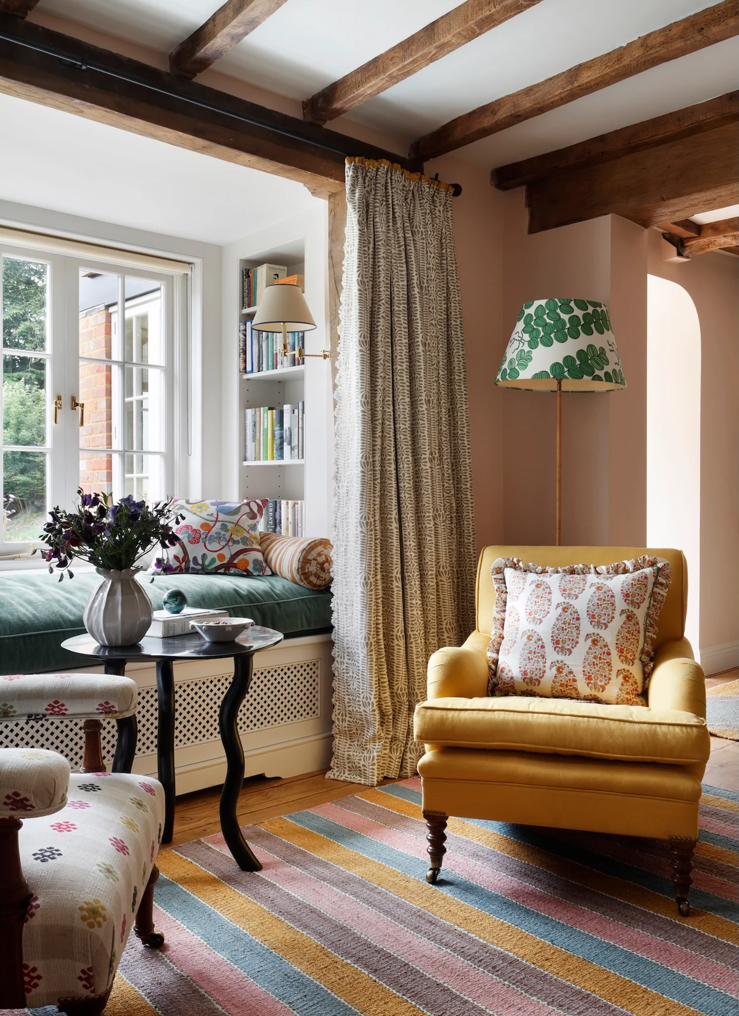 Cozy room with exposed ceiling beams, featuring a yellow armchair with a paisley pillow next to a lamp with a green patterned shade. Beside it, patterned drapes frame a window nook with a plush green seat, colorful rug, and dark side table.