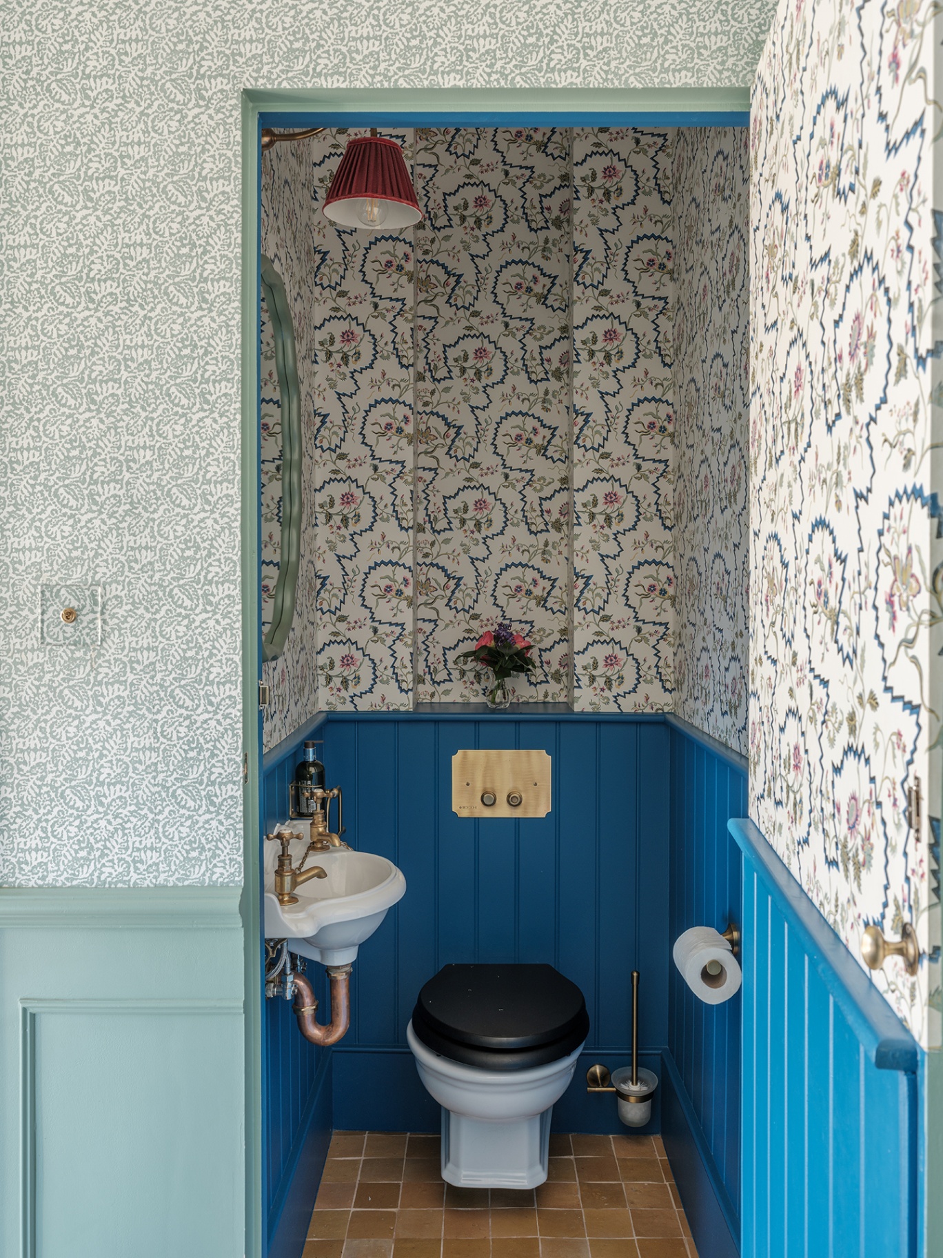 A small cloakroom with a highly patterned wallpaper on the upper half of the walls and blue painted tongue-and-groove panelling on the lower. Inside is a small wall-mounted basin and toilet, with a mirror and wall light hung on the left 