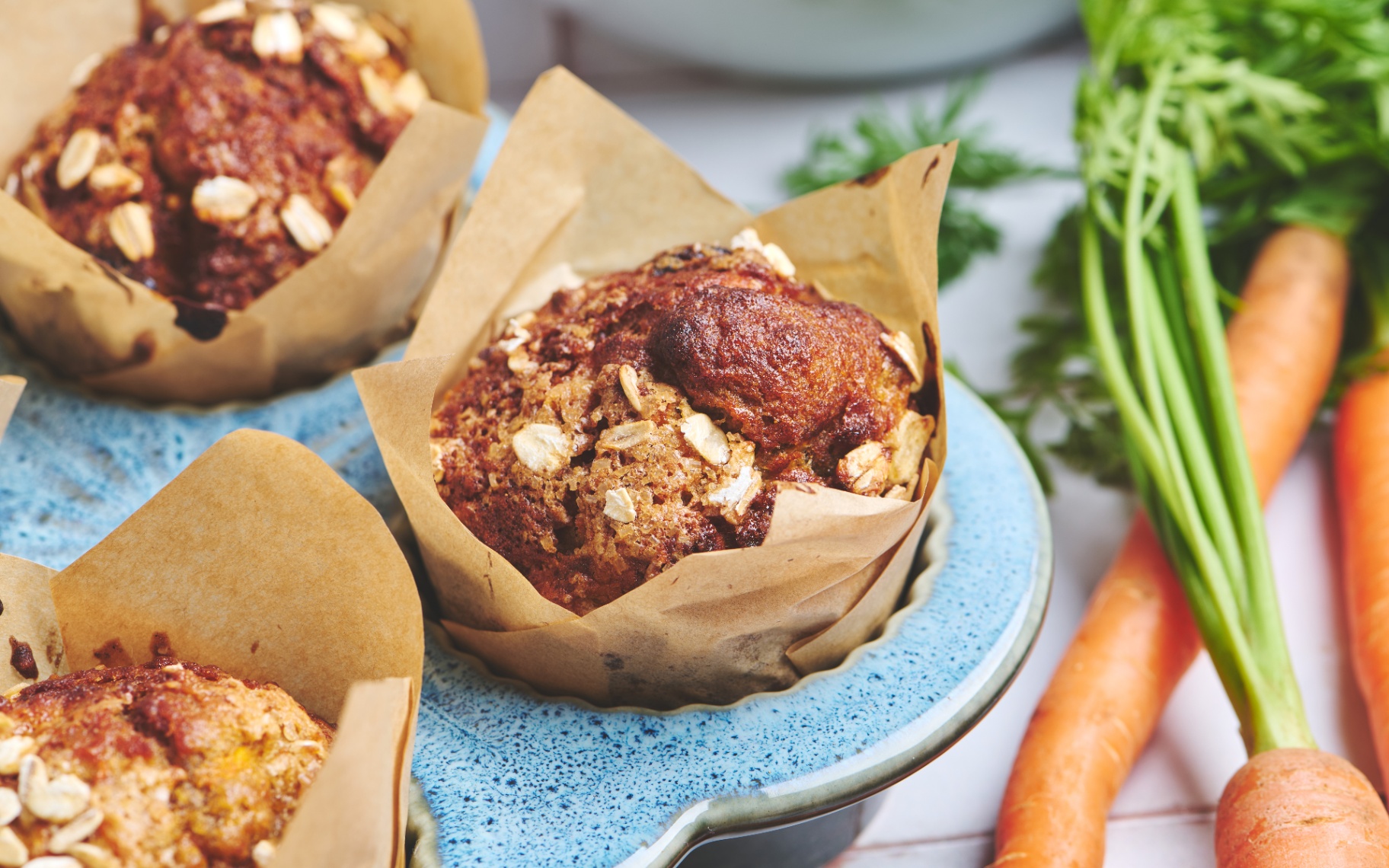 Golden-brown carrot cake breakfast muffins wrapped in brown parchment paper cases and topped with rolled oats, arranged in a blue muffin tray alongside fresh raw carrots with green leafy tops