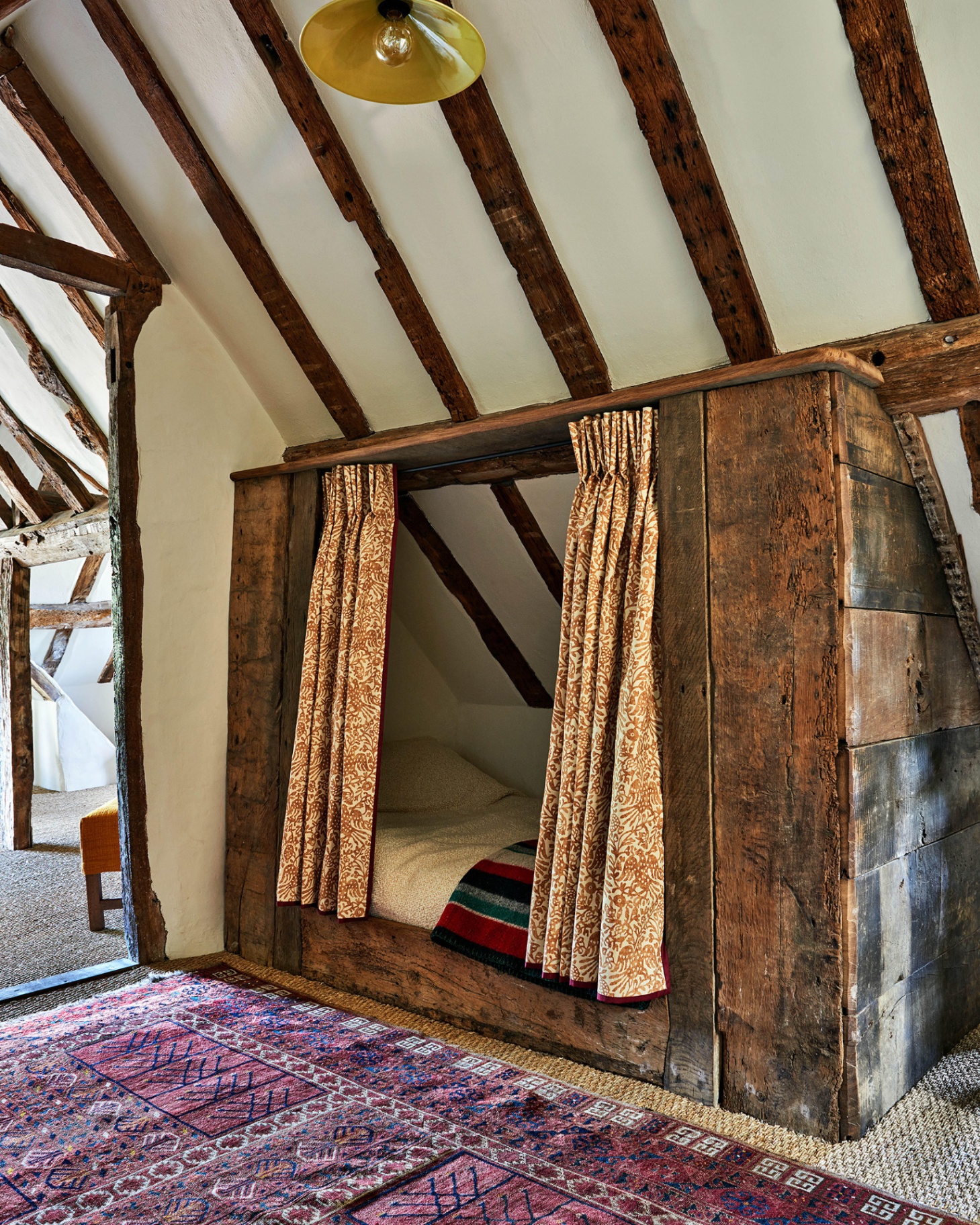 a cottage bedroom with a bed in a wooden nook, a white ceiling and walls covered in old wooden beams and a dark red persian style rug on the floor