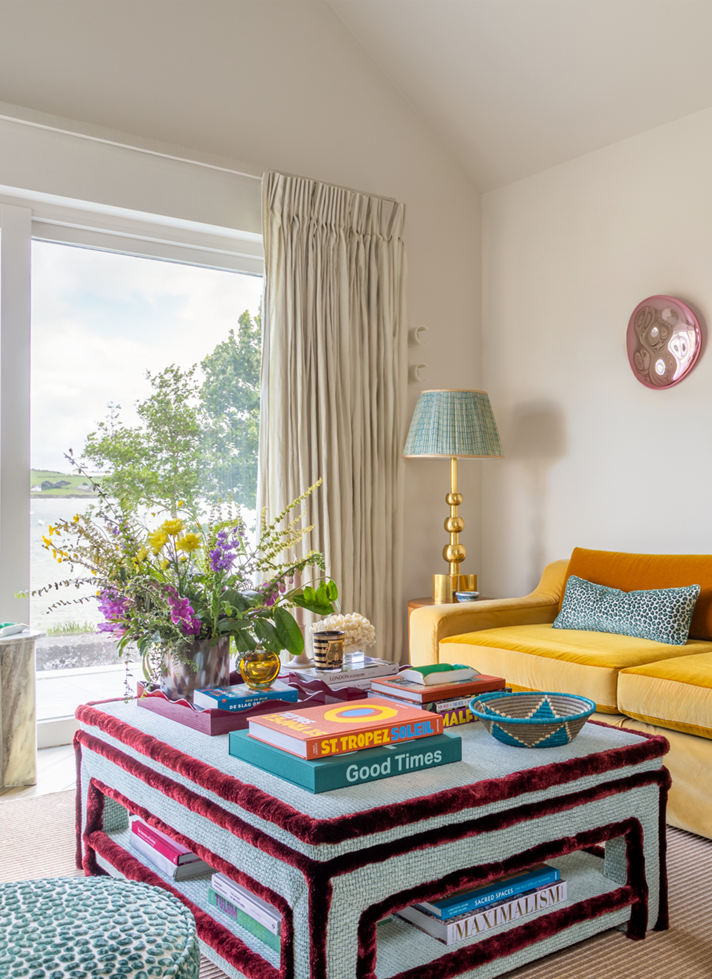 a living room with a blue and red upholstered coffee table full of books and a vase of flowers and a yellow velvet sofa behind it