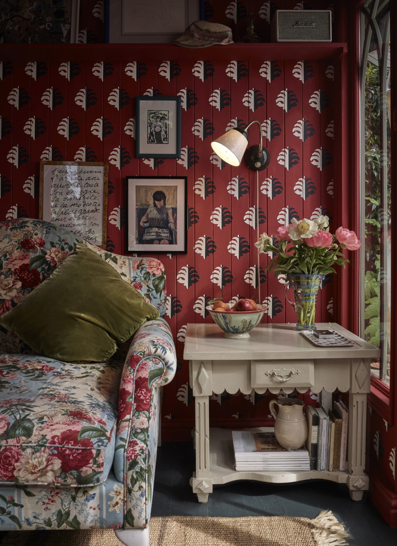 a living room corner with deep red panelled walls with black and white hand painted leaves, a blue and pink floral sofa with an olive green velvet cushion with a scalloped detail side table