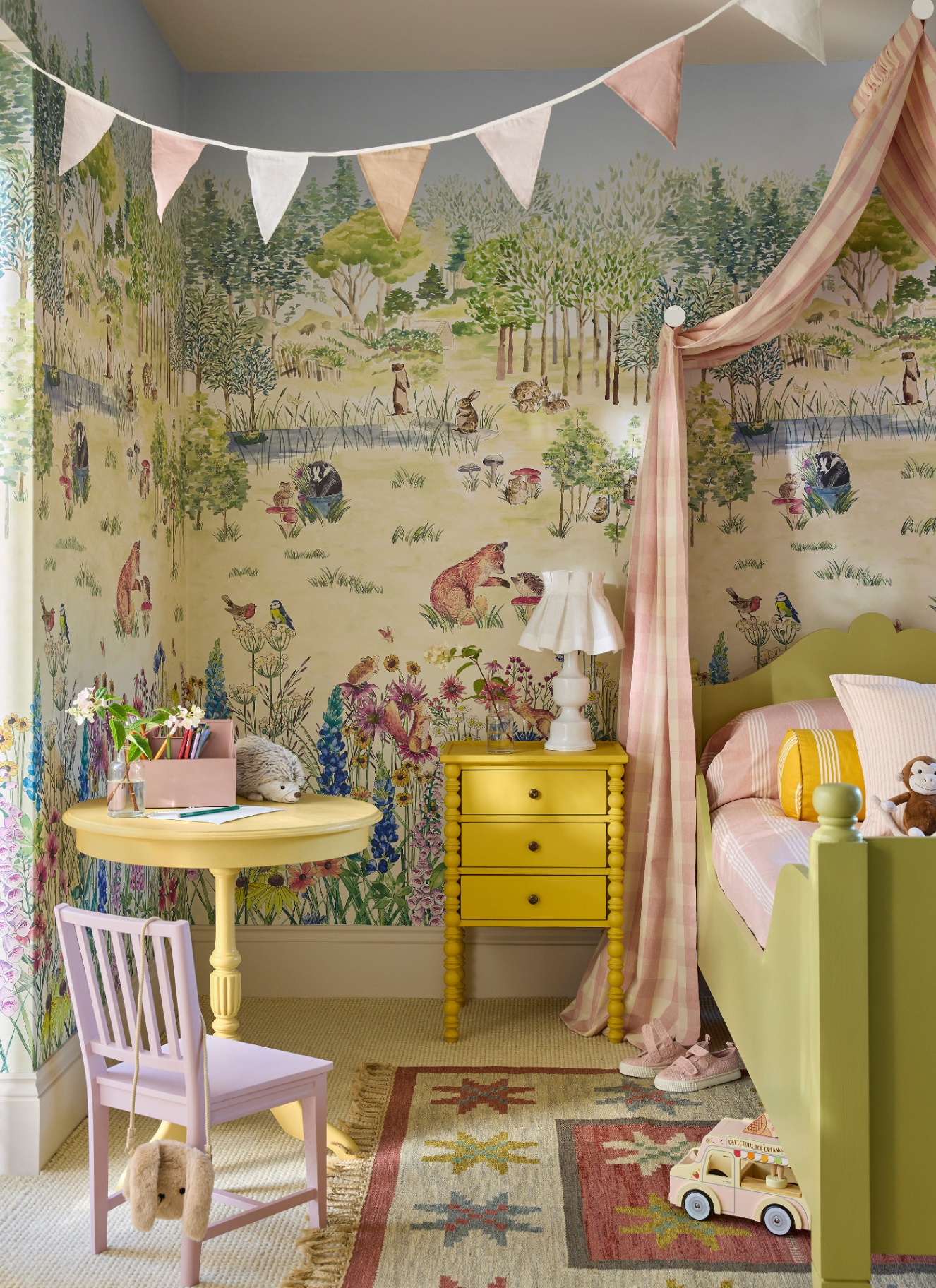 A childrens bedroom with a woodland illustrated scene wallpaper, a green bed with a pink canopy and a yellow bedside table