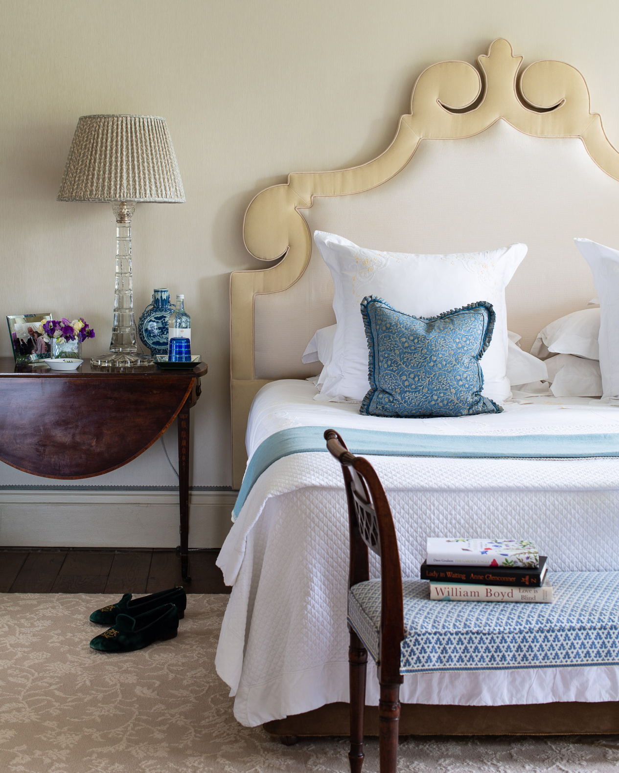 a bedroom with a big ornate cream headboard, a bed with white bedding and blue accessories, a dark wood bedside table with a large pleated lampshade on it and an upholstered bench at the end of the bed