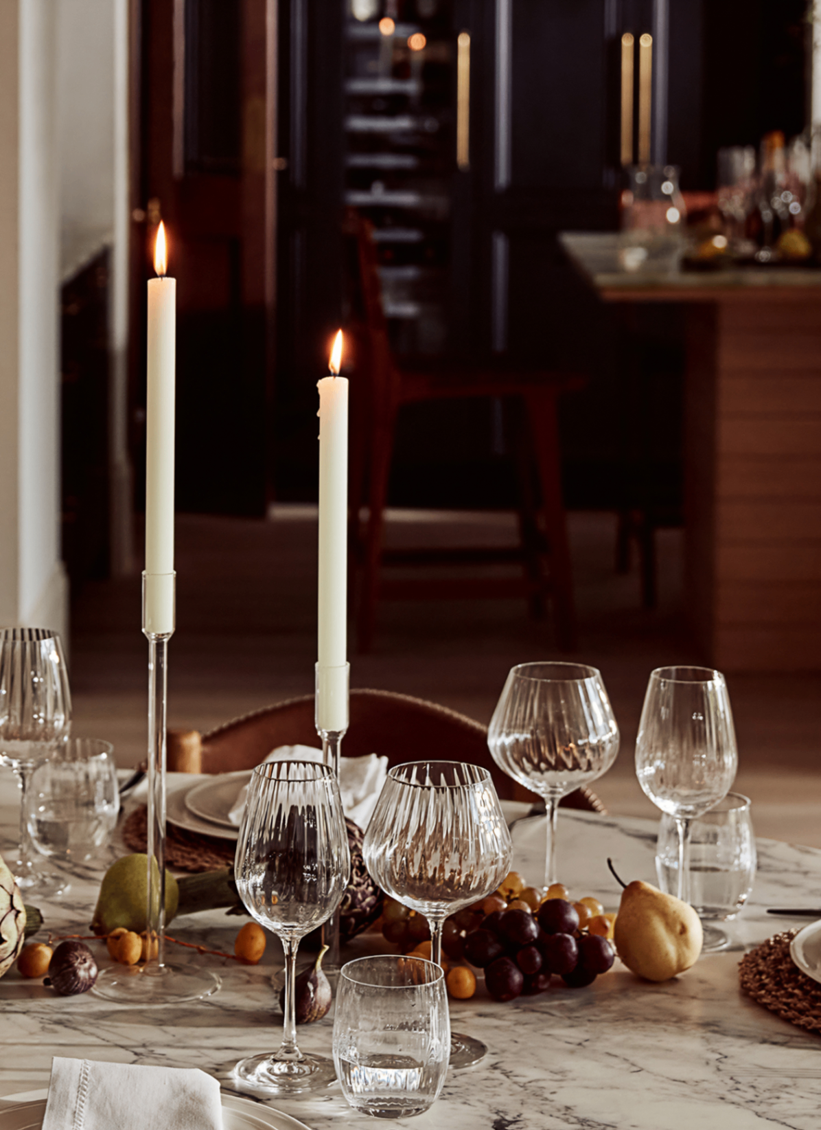 close up of a white and grey marble dining table with fresh autumnal fruit in the centre, ribbed stemmed glasses and glass candle holders with lit dinner candles