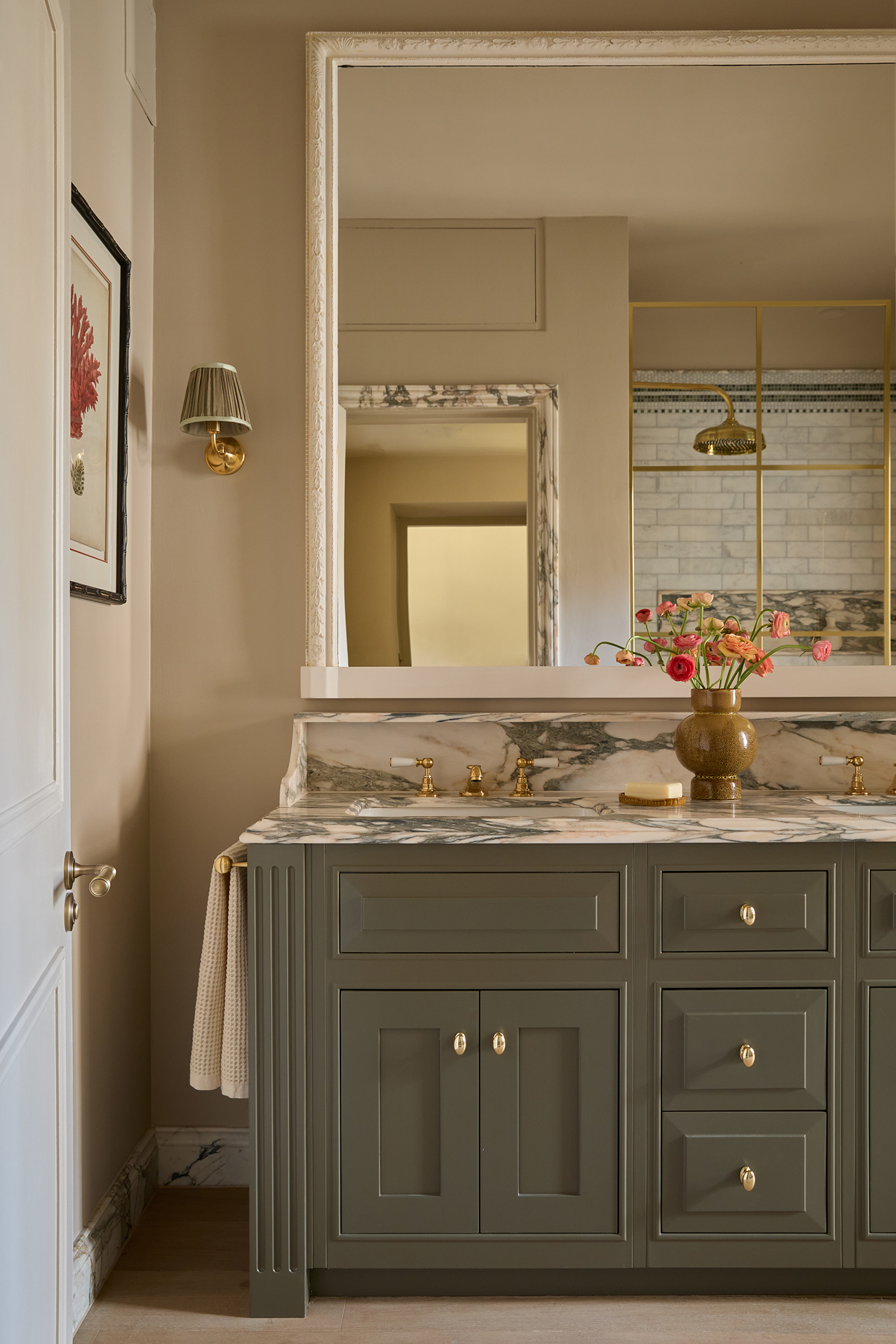 A close up of a bathroom with a green-grey vanity unit topped with marble. Above hangs a large mirror with a white frame and a small brass wall light with a pleated shade