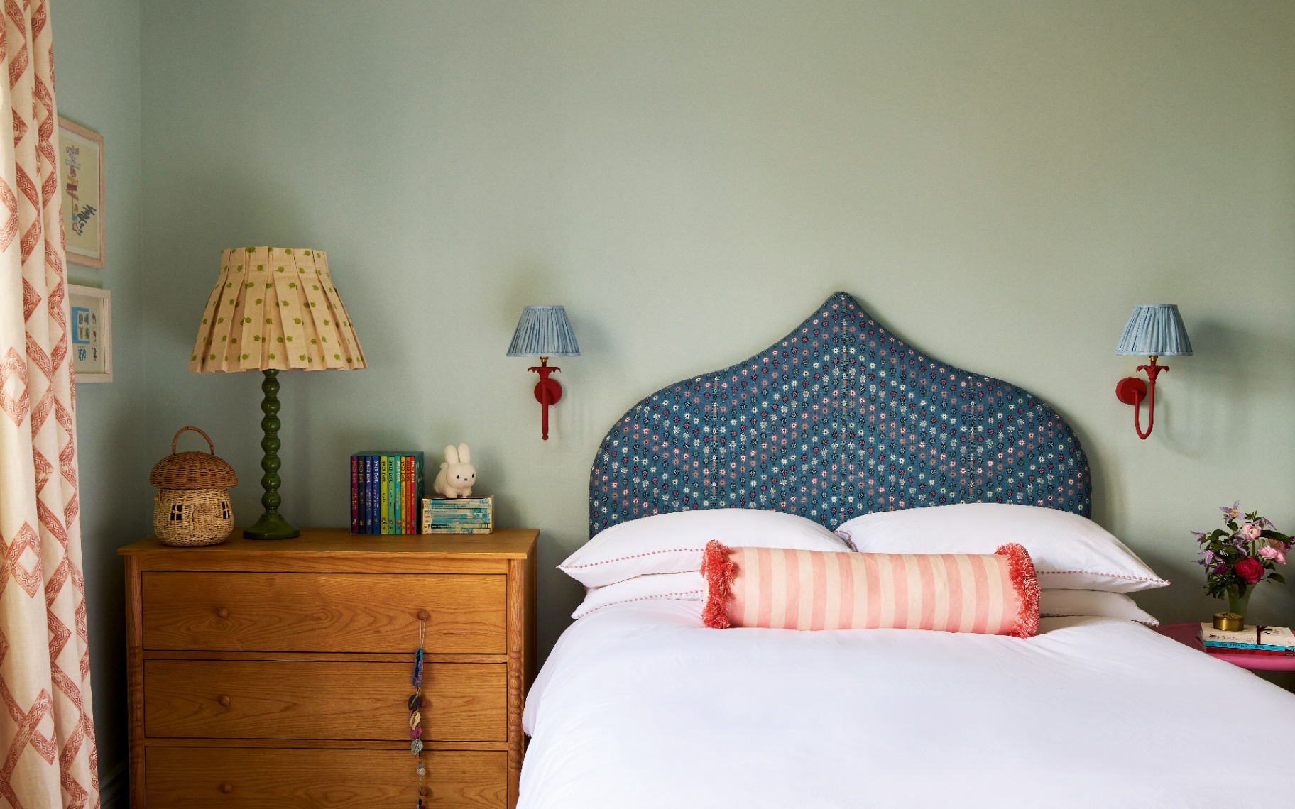 a childrens bedroom with pale green walls, a wooden chest of drawers with a pleated lamp and a stack of books and a bed with a blue patterned headboard and two red and blue sconces either side