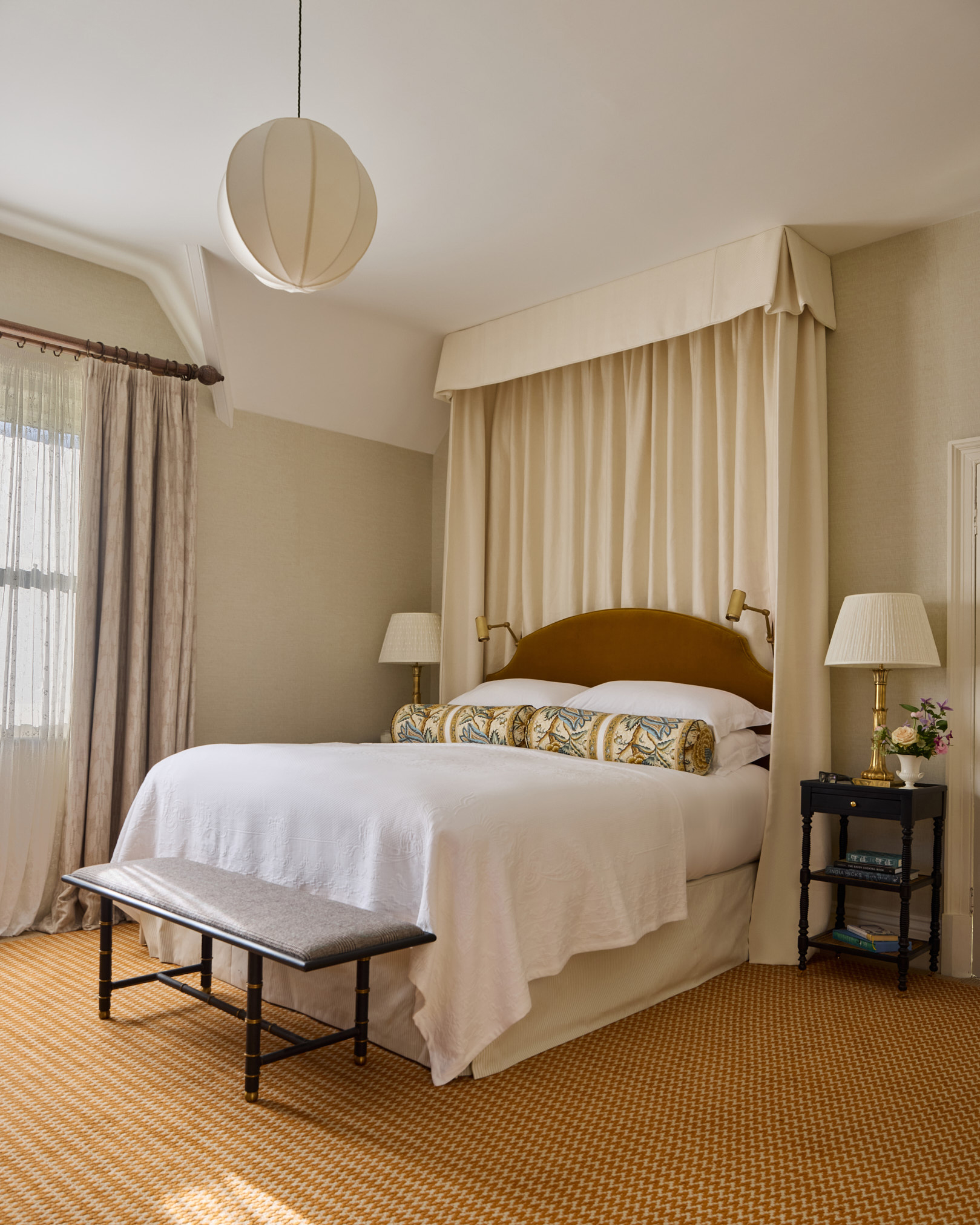 a bedroom with cream walls, a white paper sphere lampshade and a bed with a brown velvet headboard and a large cream draped canopy