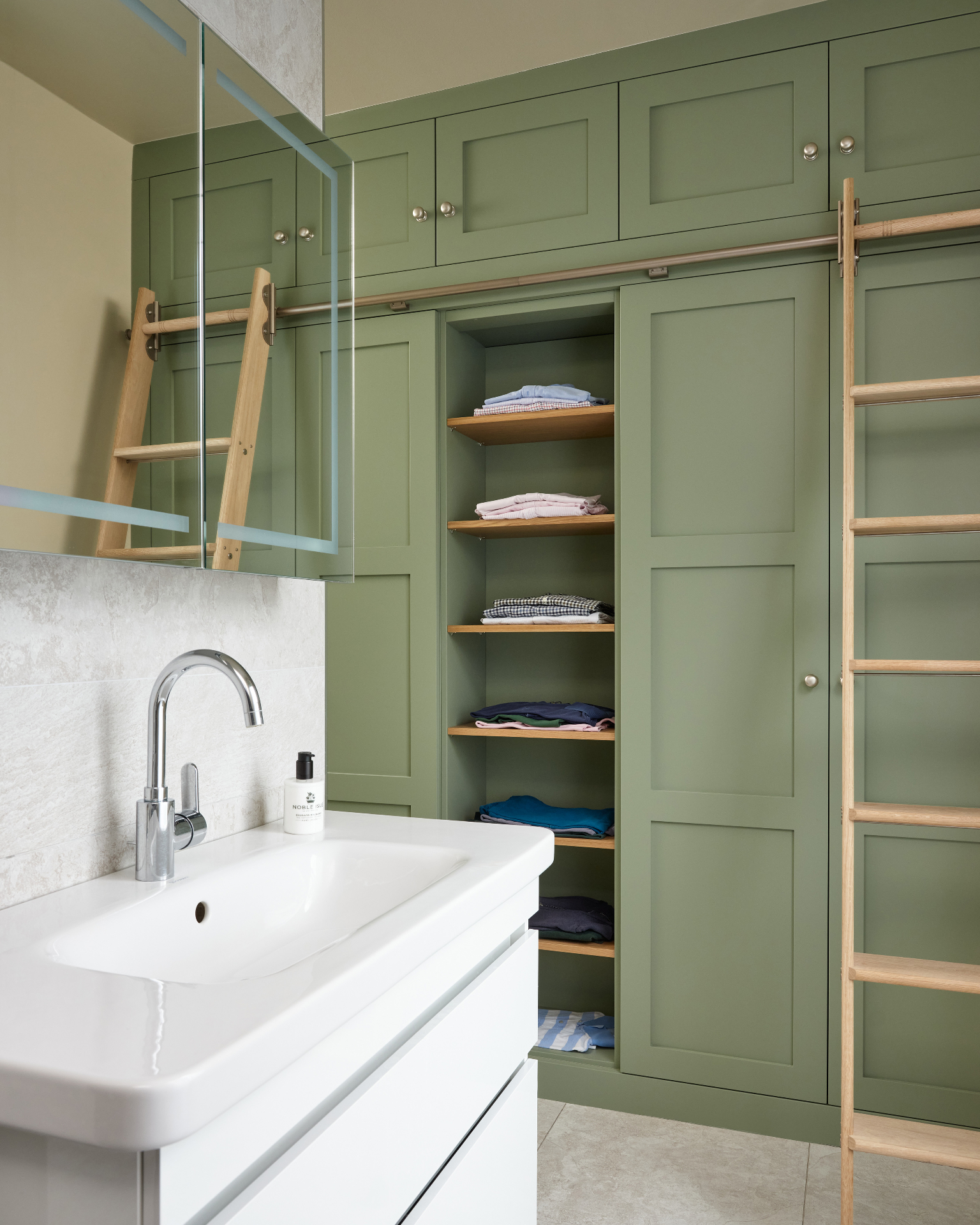a green built in dressing room in a bathroom, with vertical open shelves with folded shirts and a light wood library ladder and a white vanity unit with sink and a mirrored cabinet