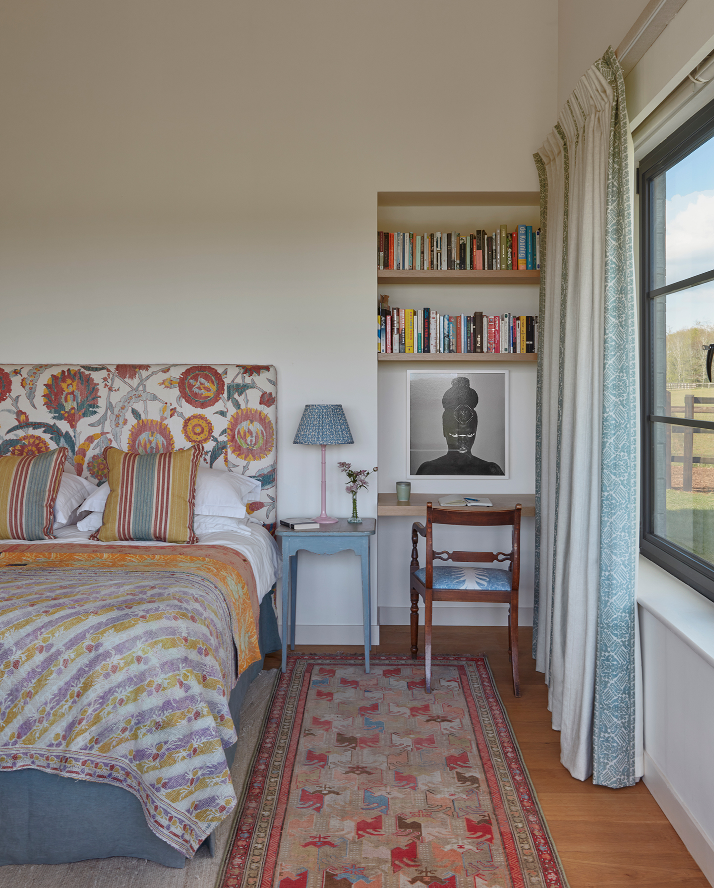 A bedroom with neutral walls. The double bed has a vibrant floral headboard and decorative cushions and throw. A blue bedside table holds a posy, book and table lamp. A built in desk is paired with a wooden chair and built-in shelves which hold books