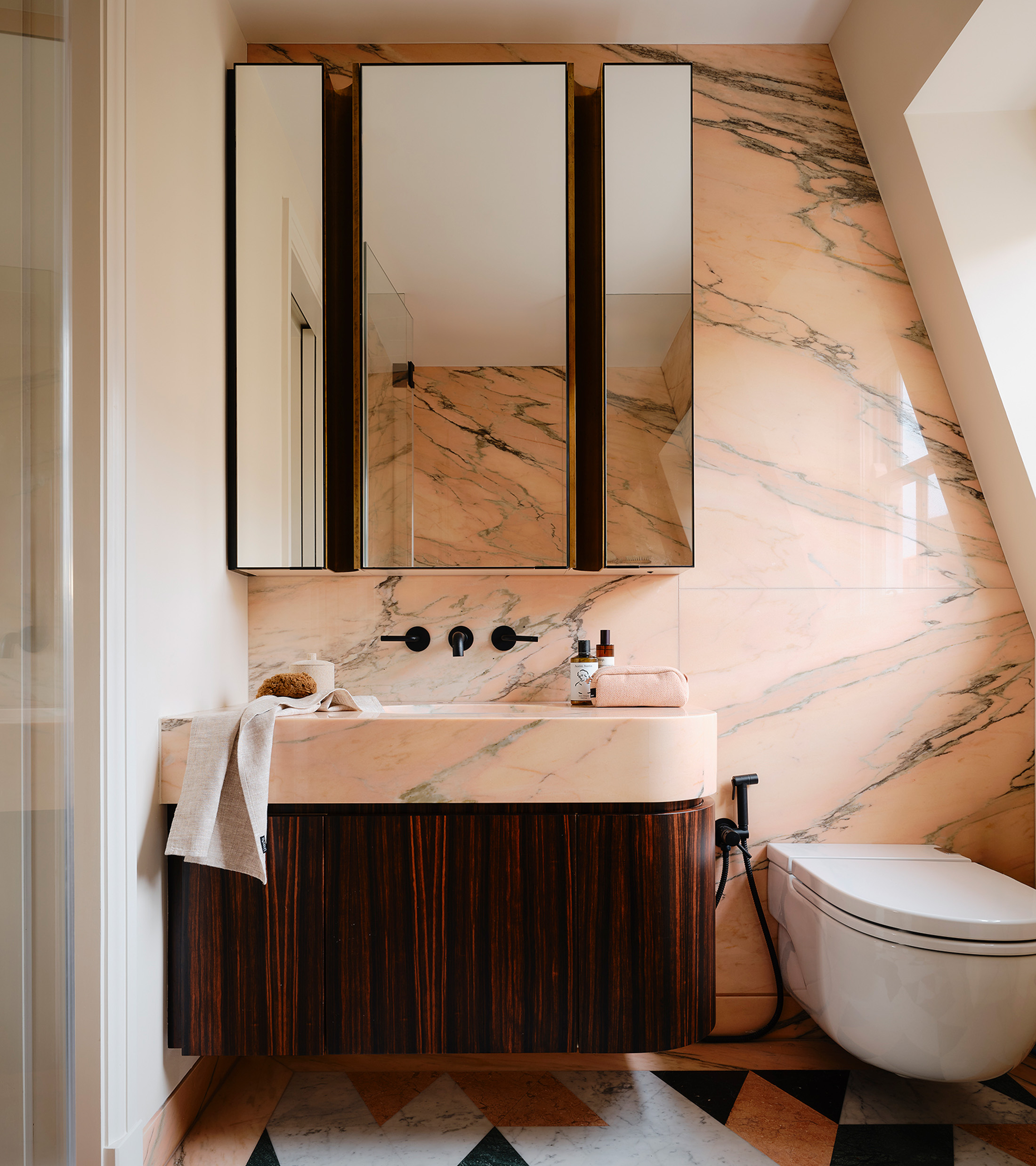 a neutral en suite with pale pink marble walls, a large mirror cabinet and a dark wood vanity