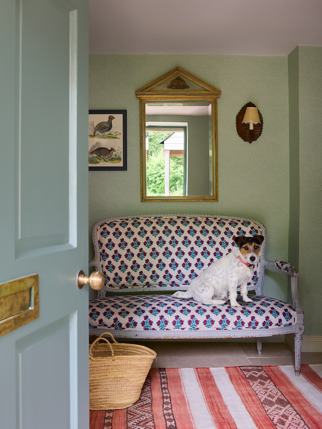 An open front door reveals an entrance hall with a striped rug, vintage bench covered in floral fabric and an ornate mirror, wall light and artwork hang above. A jack russell sits on the bench. There is a basket on the floor