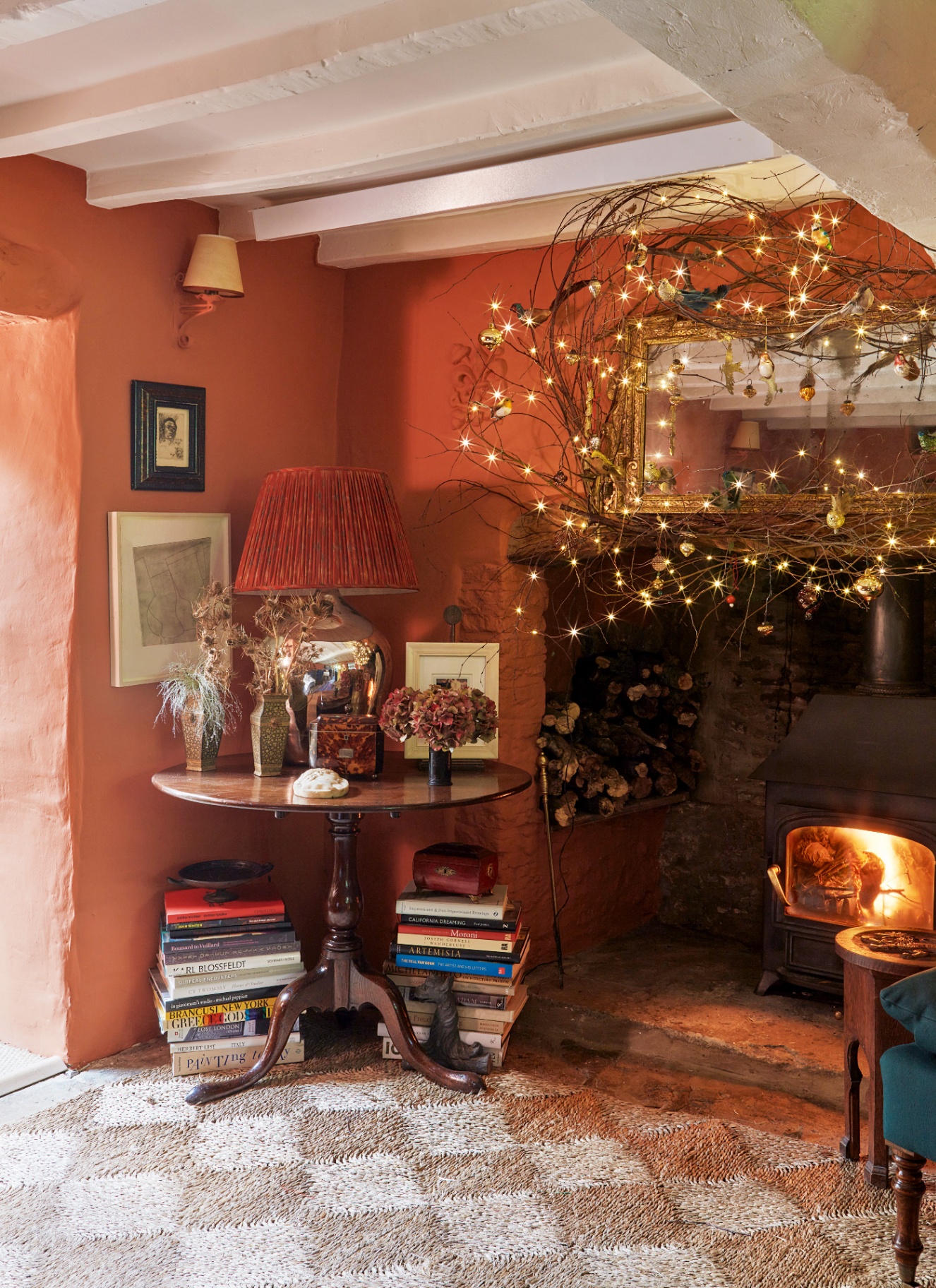 a corner of a room with terracotta walls, a round wooden table with a plated lamp and stacks of books underneath and a large fireplace with a log burner and a twig wreath above it