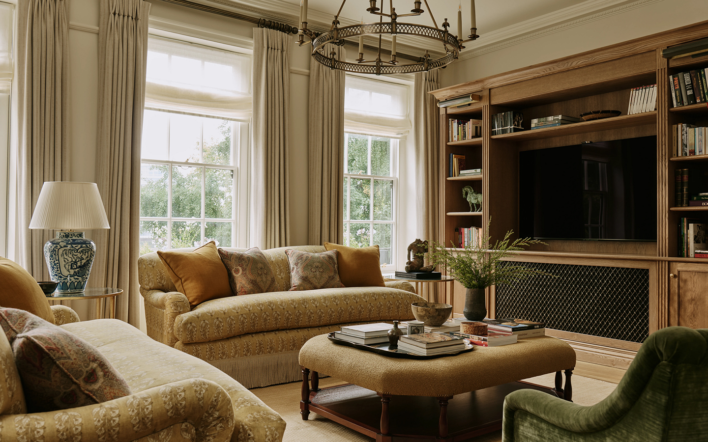 A sitting room with a pair of fringed upholstered classic sofas, green velvet chair and upholstered ottoman in the foreground. A large shelving unit holds books and a TV along the right wall.